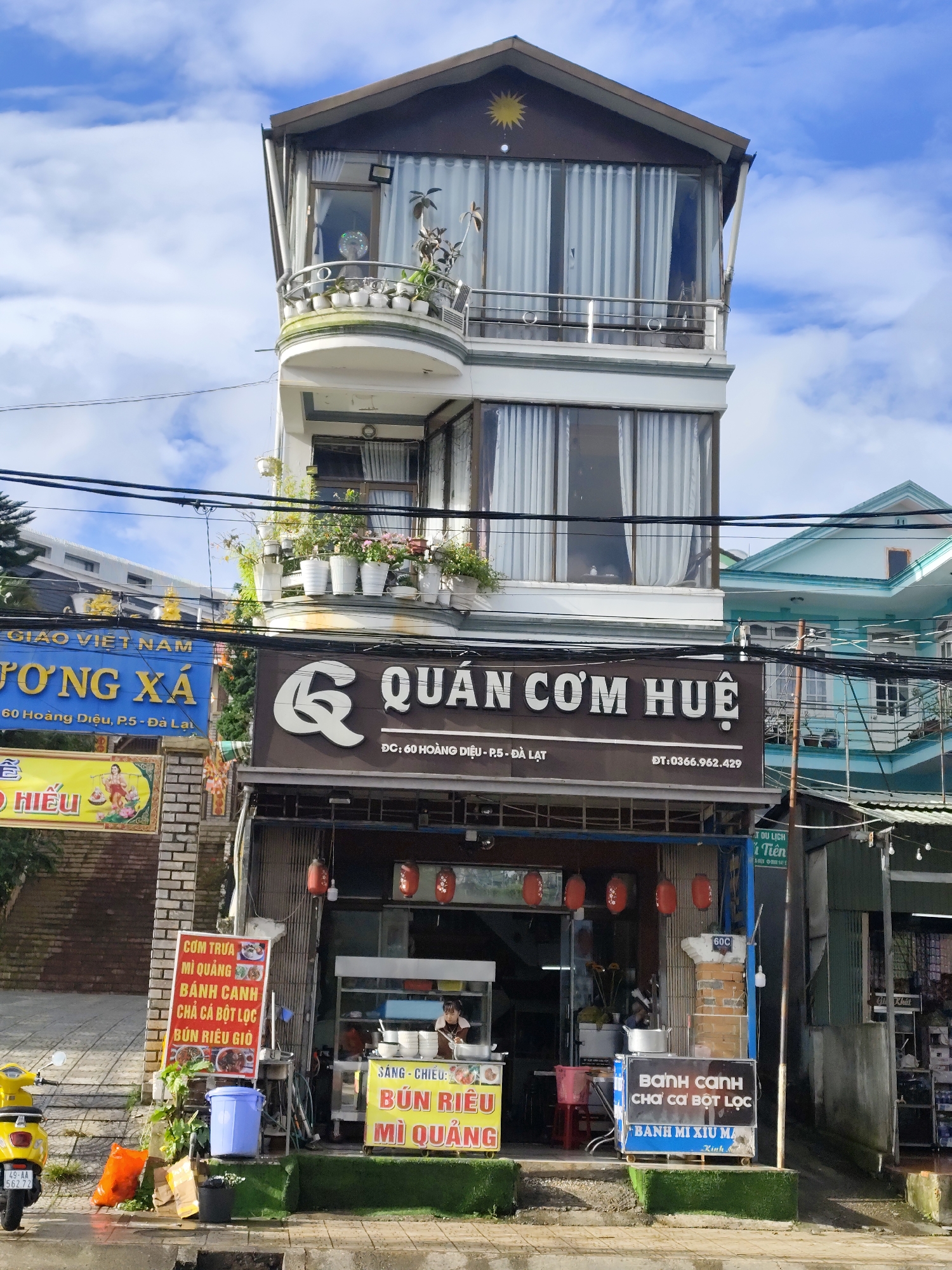 The image captures a storefront in Da Lat, Vietnam, likely a restaurant given the signage. The building is narrow and tall, with multiple stories. The ground floor features an open-fronted establishment with a counter where a woman is visible, preparing food. Red lanterns hang above the entrance. Various signs are displayed, including one with Vietnamese text and images advertising food items like "Cơm Trưa," "Mì Quảng," and "Bánh Canh." To the left, a yellow scooter is parked on the sidewalk, and a blue trash bin and some bags are also present. Above the storefront, the building has windows with white curtains and a balcony adorned with potted plants. Further up, a decorative sun motif is visible on the upper facade. The surrounding environment includes other buildings, power lines, and a bright sky with clouds. The address "60 Hoàng Diệu, P.5 - Đà Lạt" is visible on one of the signs, indicating the location.