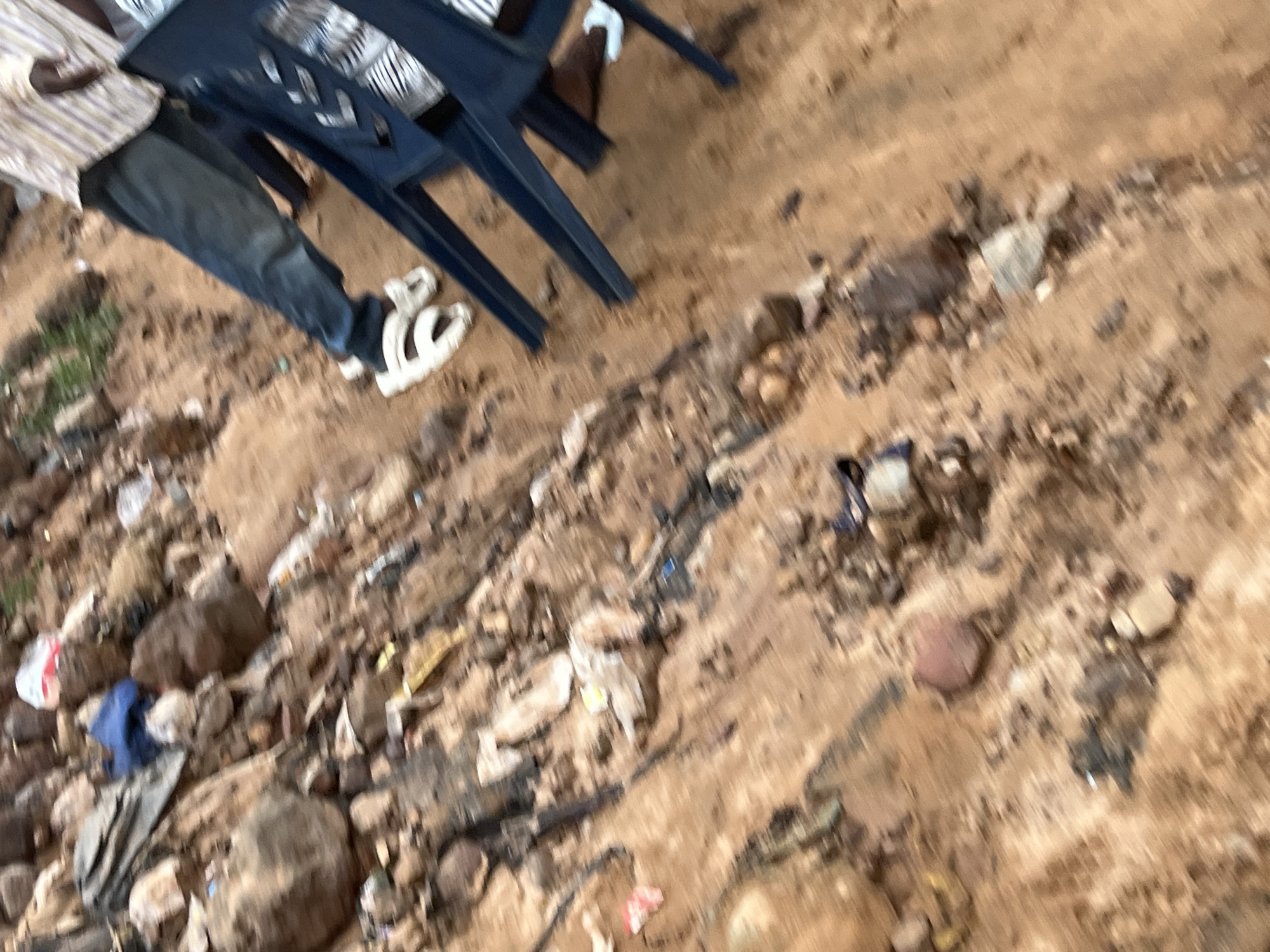 The image shows a ground-level view of a sandy, debris-strewn area. The ground is composed of dirt and small stones. Various pieces of litter, including plastic bags, wrappers, and what appear to be discarded food items, are scattered across the surface. In the upper left quadrant, a large rock is visible. To the right of the rock is a blue plastic bag, and next to that, a dark brown, rounded object that could be a rock or a piece of debris. Further to the right, a person's legs are visible wearing blue jeans and white sandals. Two dark blue plastic chairs are positioned in the middle and right portions of the image. The chairs have a slatted design. The scene appears to be outdoors, possibly in an urban or semi-urban environment with a significant amount of accumulated waste. The image is slightly blurred, suggesting motion or a quick capture.