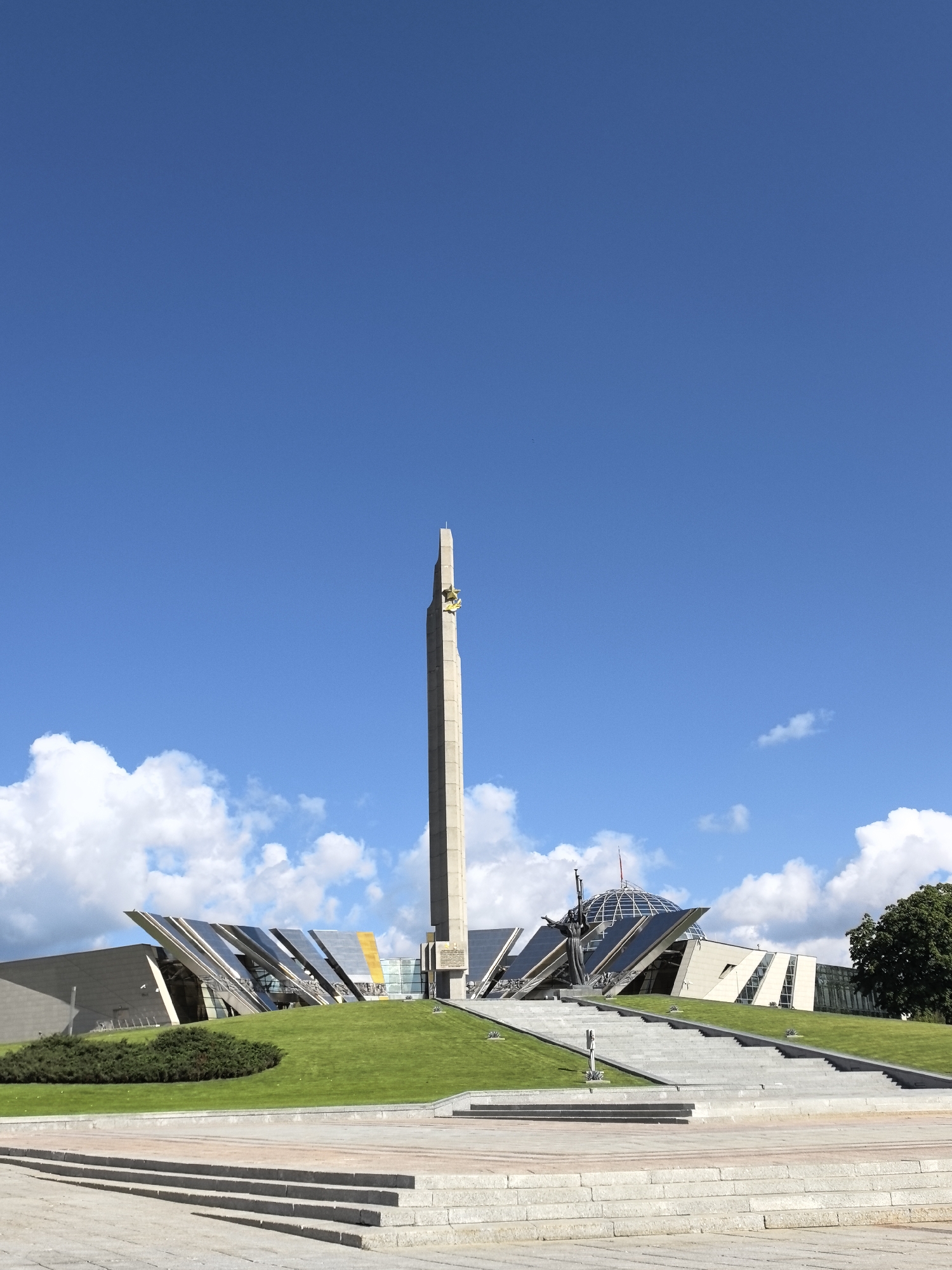 A monument stands tall against a clear blue sky with scattered white clouds. The monument is a tall, slender obelisk made of concrete, topped with a golden star. The obelisk is positioned on a grassy hill that slopes down towards a plaza with stone steps. To the left of the monument, a modern building with a unique angular roofline is visible. To the right, a large dome-shaped structure with a glass facade is connected to the modern building. A bronze statue of a figure with outstretched arms stands in front of the dome. The environment is a public space with manicured lawns and paved walkways, suggesting it is a memorial or historical site. The lighting indicates it is daytime.