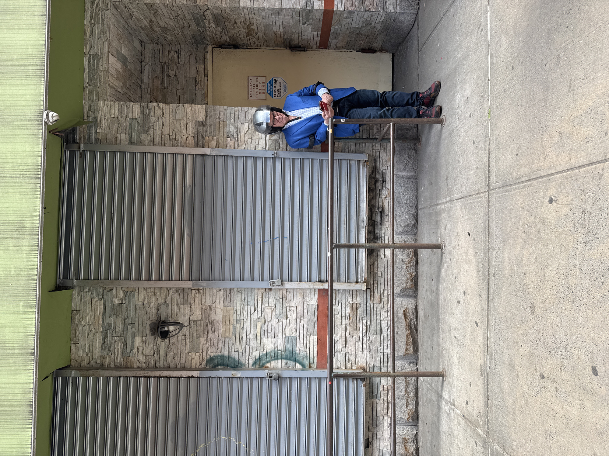 The image shows a man wearing a blue blazer and a helmet, holding a red object, laying on metal bars next to a closed metal door in an alleyway. The walls are made of stone and there's a closed yellow door in the background with signs. The ground is concrete. The location is assumed to be New York City based on the prompt.