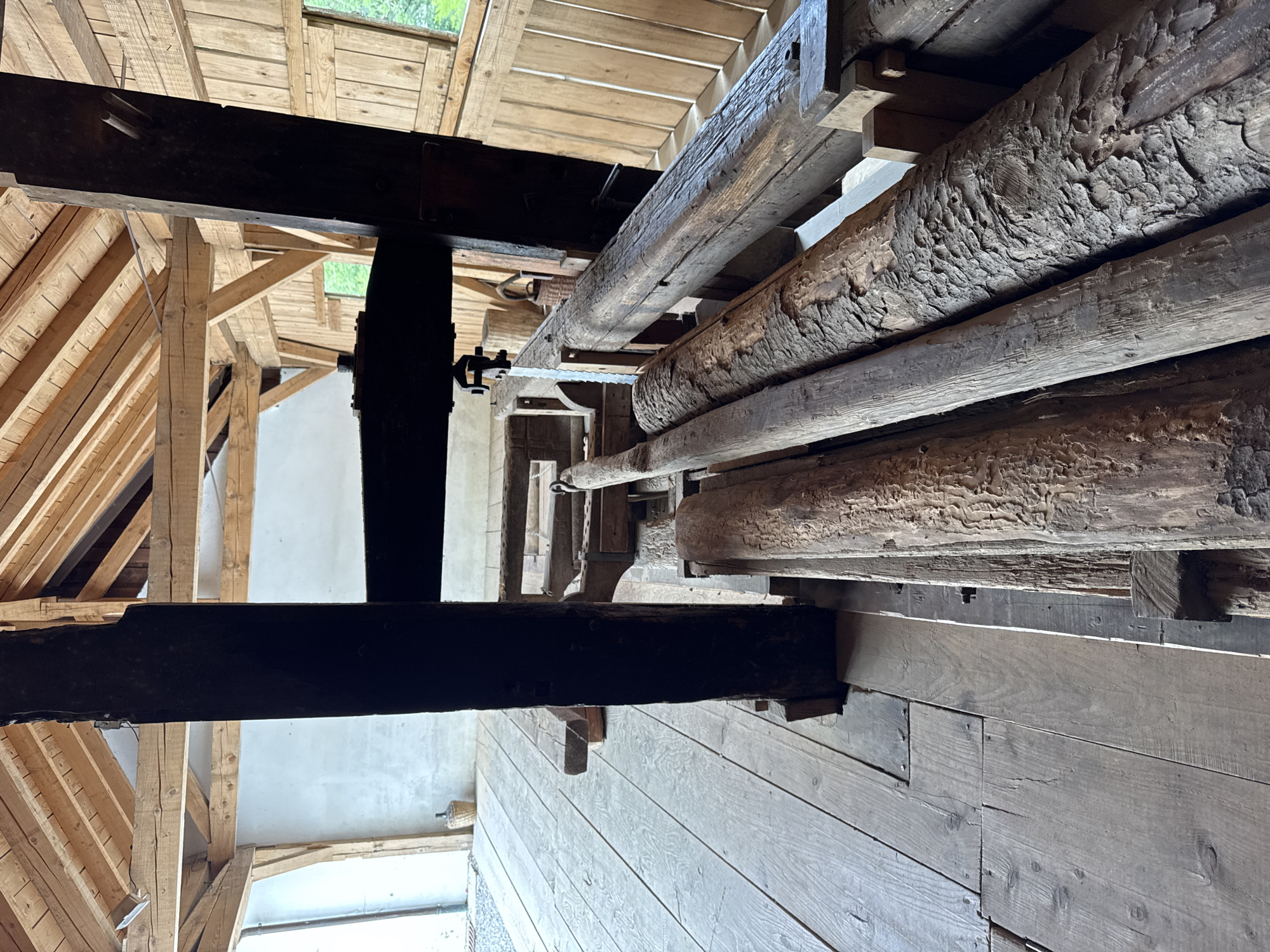 The image depicts the interior of a rustic, wooden structure, likely a historical mill or workshop, located in Postojna, Slovenia. The scene is dominated by heavy wooden beams, logs, and a wooden floor. In the foreground, several large, rough-hewn logs are positioned horizontally, their bark textured and weathered. These logs rest on a wooden platform or base.
Behind the logs, a complex wooden mechanism is visible, consisting of beams, supports, and what appears to be a large saw blade. This machinery is integrated with thick, dark wooden vertical support posts.
The floor is constructed from wide, weathered wooden planks laid lengthwise. The overall impression is one of age and functional utility. The background reveals more wooden structures, including rafters and walls made of planks. Natural light enters from windows or openings, casting shadows and highlighting the textures of the wood. There are no people visible in the image. The environment suggests a place dedicated to woodworking or processing timber.