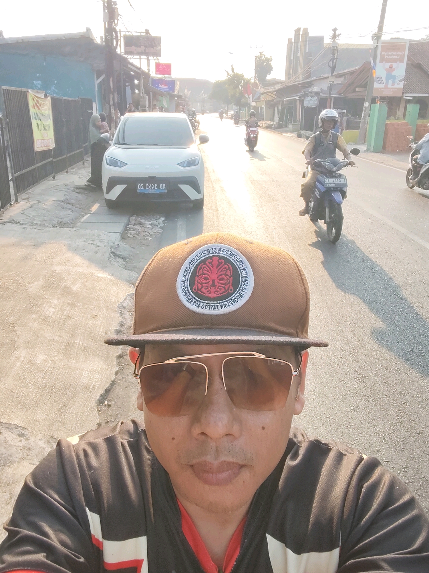 The image is a selfie taken outdoors in Purwakarta, Indonesia, during daylight. In the foreground, a person wearing a brown baseball cap and sunglasses is visible, partially obscuring their face and shoulders. They are also wearing a black jacket with red and white accents. The background shows a street scene with several vehicles, including a white car and multiple motorcycles. One motorcycle is ridden by a person wearing a helmet and a yellow shirt, while another motorcycle is parked in the distance. Buildings and power lines line the street, and banners are displayed on some of the buildings. The overall atmosphere suggests a typical day in a bustling urban environment.
