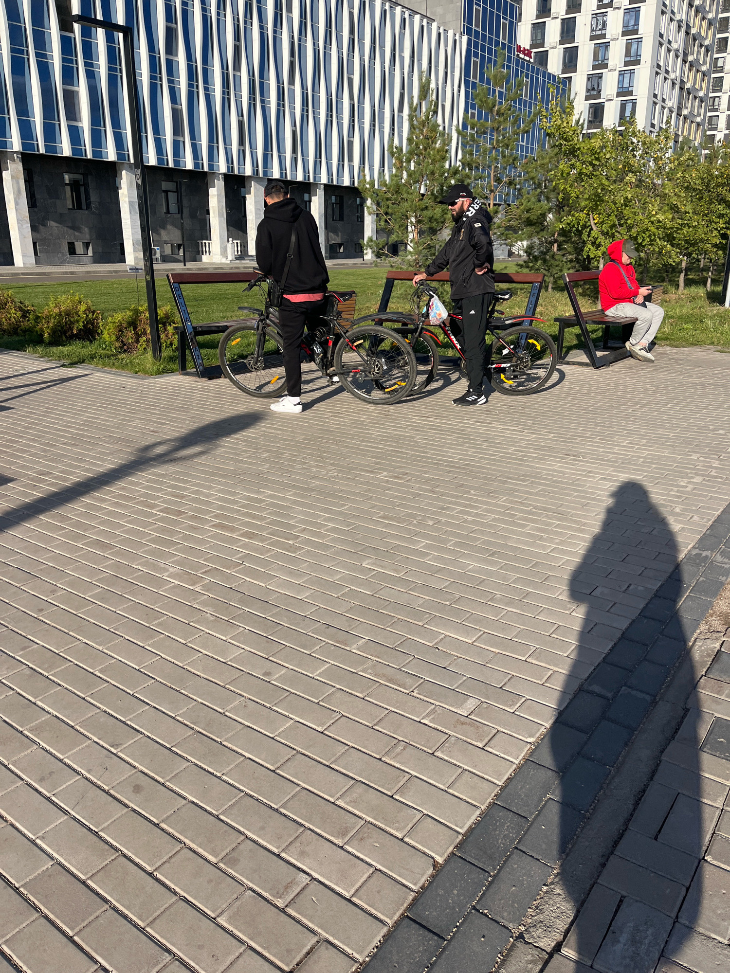 The image depicts an outdoor scene in a modern urban setting, likely in Astana, Kazakhstan, given the architectural style of the buildings. In the foreground, a paved walkway made of rectangular grey bricks is visible, with a shadow of a person cast across it. Two men are standing near bicycles, with one man wearing a black hoodie and black pants, and the other in a black jacket and black pants. A third person, wearing a red hoodie and grey pants, is sitting on a bench in the background. The background features contemporary buildings with distinctive facades, including a building with alternating blue and white vertical elements and another with a grid of windows. Trees and green shrubbery are also present, indicating a park or landscaped area adjacent to the buildings. The overall impression is a sunny day in a well-maintained urban environment.