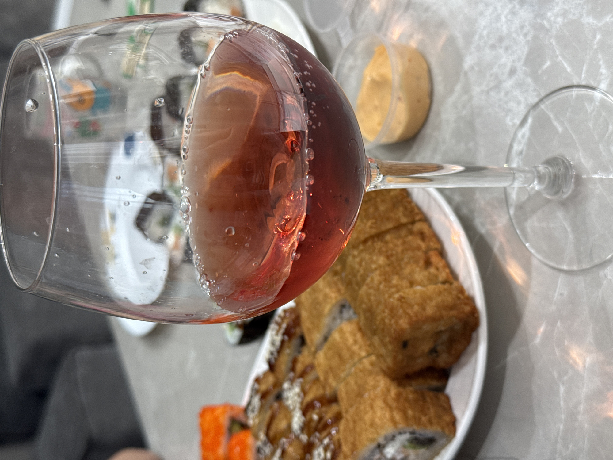An indoor scene captures a close-up view of a dining arrangement, featuring a wine glass and various food items. The dominant subject is a clear, stemmed wine glass positioned in the foreground, filled with a reddish-pink liquid, likely rosé wine. Numerous small bubbles are visible clinging to the inside surface of the glass, indicating effervescence.
In the mid-ground and background, several plates of food are present. One prominent plate holds multiple pieces of deep-fried sushi rolls, characterized by their golden-brown, crispy exterior. Some of these rolls are topped with a light-colored, creamy sauce. Another partially visible plate shows more traditional sushi rolls, clearly identifiable by vibrant orange roe (possibly tobiko or masago). A small, clear plastic container filled with a creamy, yellowish-orange dipping sauce is also seen.
The food and drink are arranged on a light-colored table with a subtle, possibly marble or stone pattern. The background is softly blurred, showing indistinct shapes that suggest other furniture or elements within the room. The overall ambiance is casual, indicative of a relaxed meal or snack. The lighting appears to be soft and ambient, consistent with an indoor setting. No discernible text is visible within the image. This scene takes place in Murom, Russia.