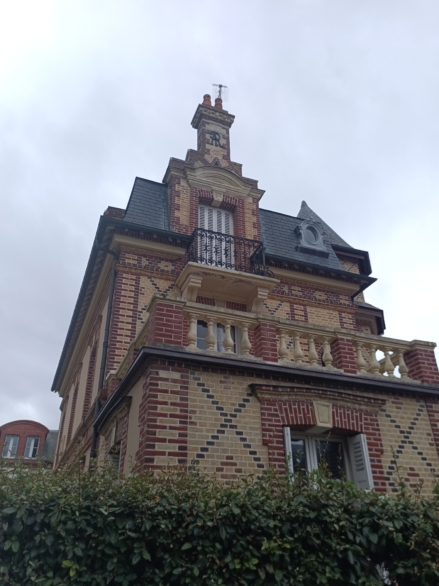 This is a low-angle, eye-level shot of a brick building in Houlgate, France, on a cloudy day. The building features a complex architectural design with red and tan bricks laid in a patterned manner. The main focus is on a section of the building with a decorative balcony, a balustrade, and a dormer window with a circular opening. A chimney with what appears to be an antenna on top is visible against the overcast sky. In the foreground, a dense green hedge runs across the bottom of the frame, obscuring the base of the building. The image captures architectural details such as decorative brickwork, wrought-iron railings, and various window styles. No people or animals are visible in the scene.