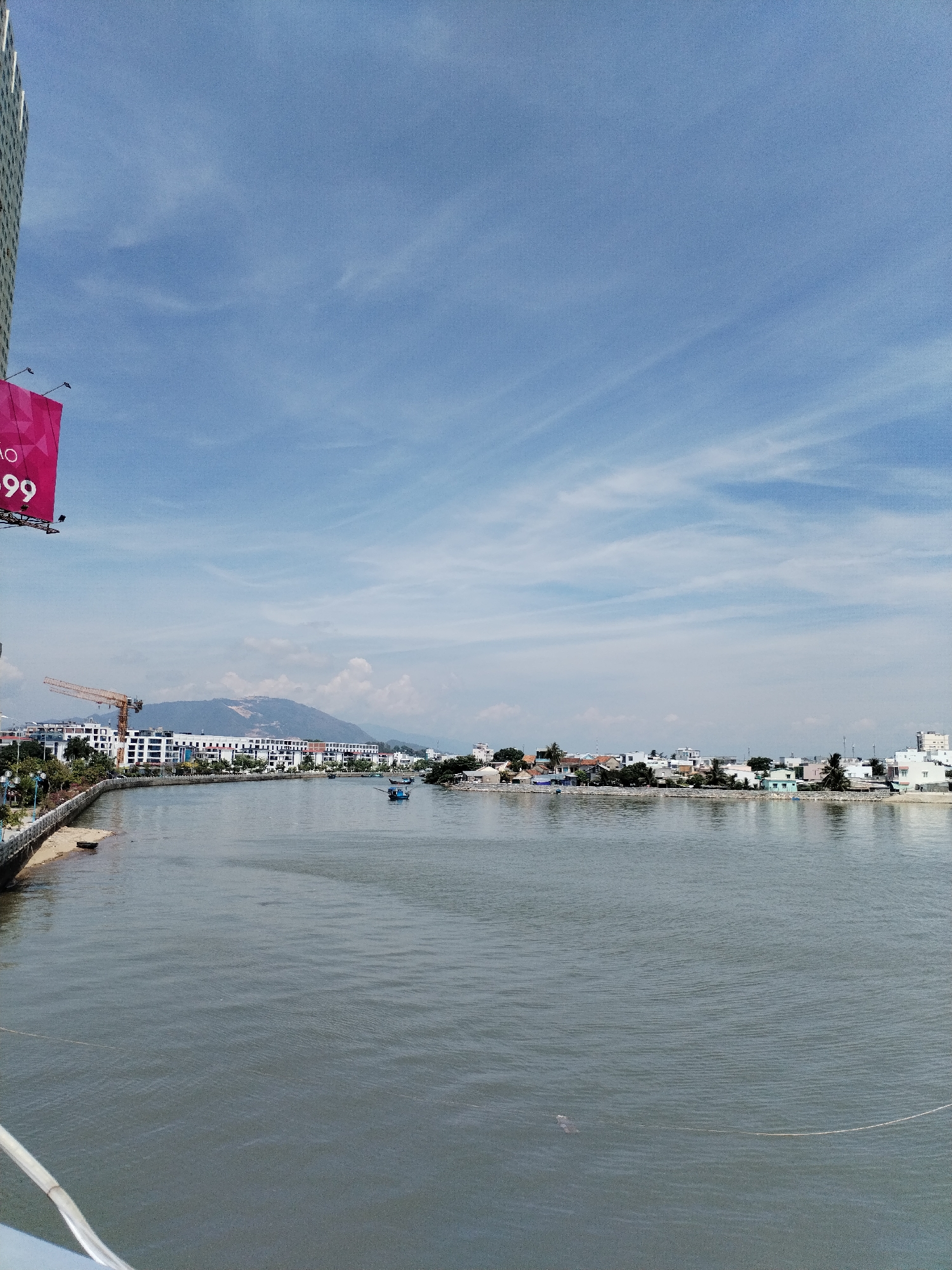 This media file captures a scenic view of a river running through a coastal city in Bac Nha Trang Ward, Vietnam. The scene is bathed in daylight, indicated by the bright sky and clear visibility. The weather appears pleasant, with a blue sky dotted with scattered white clouds, suggesting a sunny day.
The primary subjects include a wide river in the foreground, reflecting the sky and the surrounding landscape. On the far bank of the river, a line of buildings, varying in size and architectural style, stretches along the coastline, interspersed with lush green trees. Some of these buildings appear to be residential homes with white or light-colored walls, while others are larger, possibly commercial establishments or hotels. A prominent yellow construction crane is visible on the left side of the riverbank, hinting at ongoing development in the area.
Further down the river, several small boats are scattered, some appearing to be fishing vessels, indicating a maritime or fishing community. In the background, a large, hazy mountain range rises majestically, providing a natural backdrop to the urban landscape. The water itself is calm, with gentle ripples on its surface.
On the left side of the image, a portion of a tall, modern building is visible, suggesting a more developed urban area. Also on the left, a large, vibrant pink billboard is partially visible, displaying white text. The visible text reads "AO" and "99," possibly part of a larger advertisement.
The overall impression is one of a bustling yet serene waterside community, where nature and urban development coexist. The scene evokes a sense of everyday life in a Vietnamese coastal city, with elements of industry, residence, and natural beauty. There are no discernible people in the image, but their presence is implied by the occupied buildings and the boats on the water.