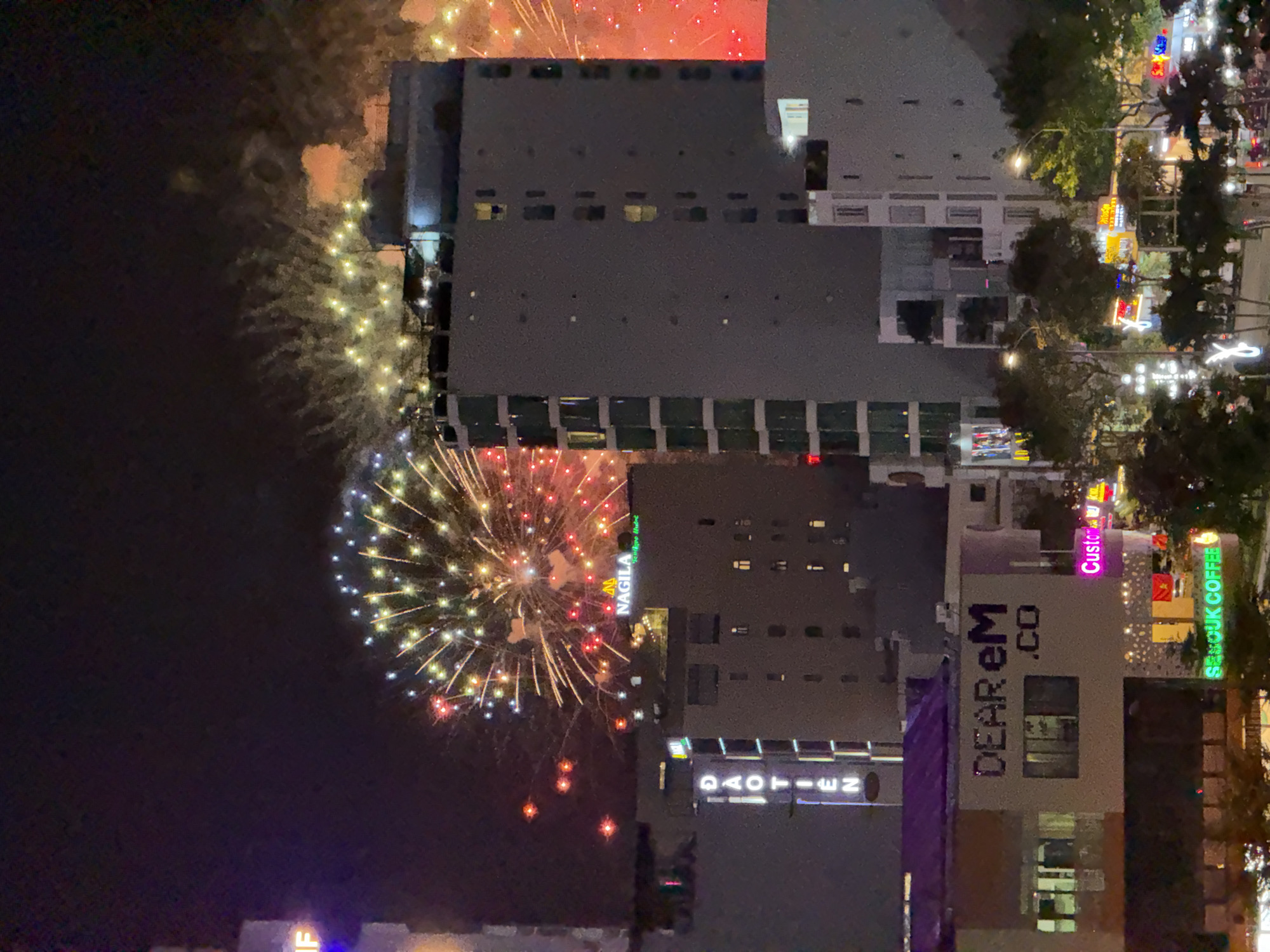 The image captures a nighttime scene in the city of Đà Nẵng, Vietnam, with a prominent display of fireworks illuminating the sky. In the foreground, several buildings of varying heights are visible. One building on the left features vertical neon lights spelling out "ĐAO TIÊN". Another building in the center has a sign that reads "NAGILA". Below this, a sign says "DEAR eM .CO". Further down, a brightly lit sign displays "SENGOUK COFFEE". The lower portion of the image shows trees and streetlights, suggesting a street-level view or a vantage point from a higher floor overlooking a busy area. The overall atmosphere is festive, with the vibrant fireworks suggesting a celebration or event.
