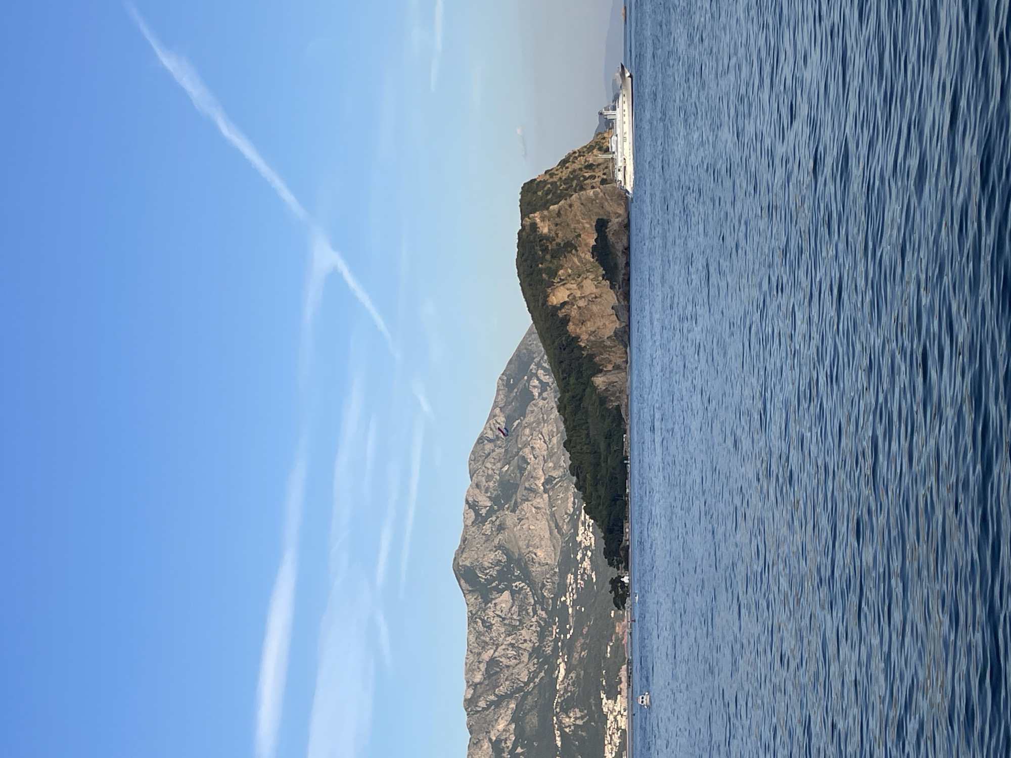 The image shows a coastal scene with a body of water in the foreground, a rocky island with trees in the middle ground, and mountains with buildings in the background. The sky is blue with some wispy clouds.
In the foreground, the water is a deep blue with gentle ripples. A white yacht is visible in the middle ground, to the right of the island. The island is covered in dense green trees, with a sheer, rocky cliff face on its seaward side. In the background, a range of rugged mountains dominates the horizon. Several buildings, seemingly houses and smaller structures, are scattered across the lower slopes of the mountains. The overall impression is one of a serene, natural landscape with a touch of human settlement.
The scene is likely the coast near Budva, Montenegro, given the distinctive mountainous coastline and the presence of islands and the sea. The Sveti Stefan island, famous for its historic fortified village, is a prominent feature of the Budva Riviera and could be the island depicted in the image, though its distinctive buildings are not clearly visible from this perspective. The presence of the yacht suggests it is a popular tourist area.