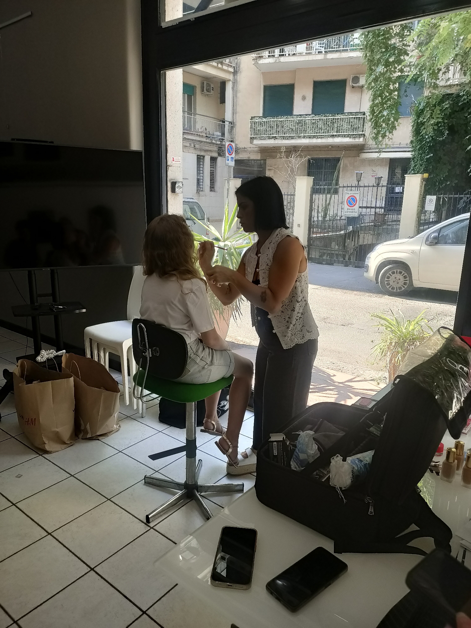 A person with shoulder-length blonde hair is seated on a green-seated swivel chair, facing away from the camera. A person with dark hair and a white lacy top is applying makeup to the seated person's face with a brush. The background shows a street scene with buildings, a white car, and lush green plants. In the foreground, there is a large open black makeup bag with various items inside, two brown shopping bags, and two cell phones on a white table. The scene appears to be taking place indoors, near a large window or doorway that looks out onto the street.