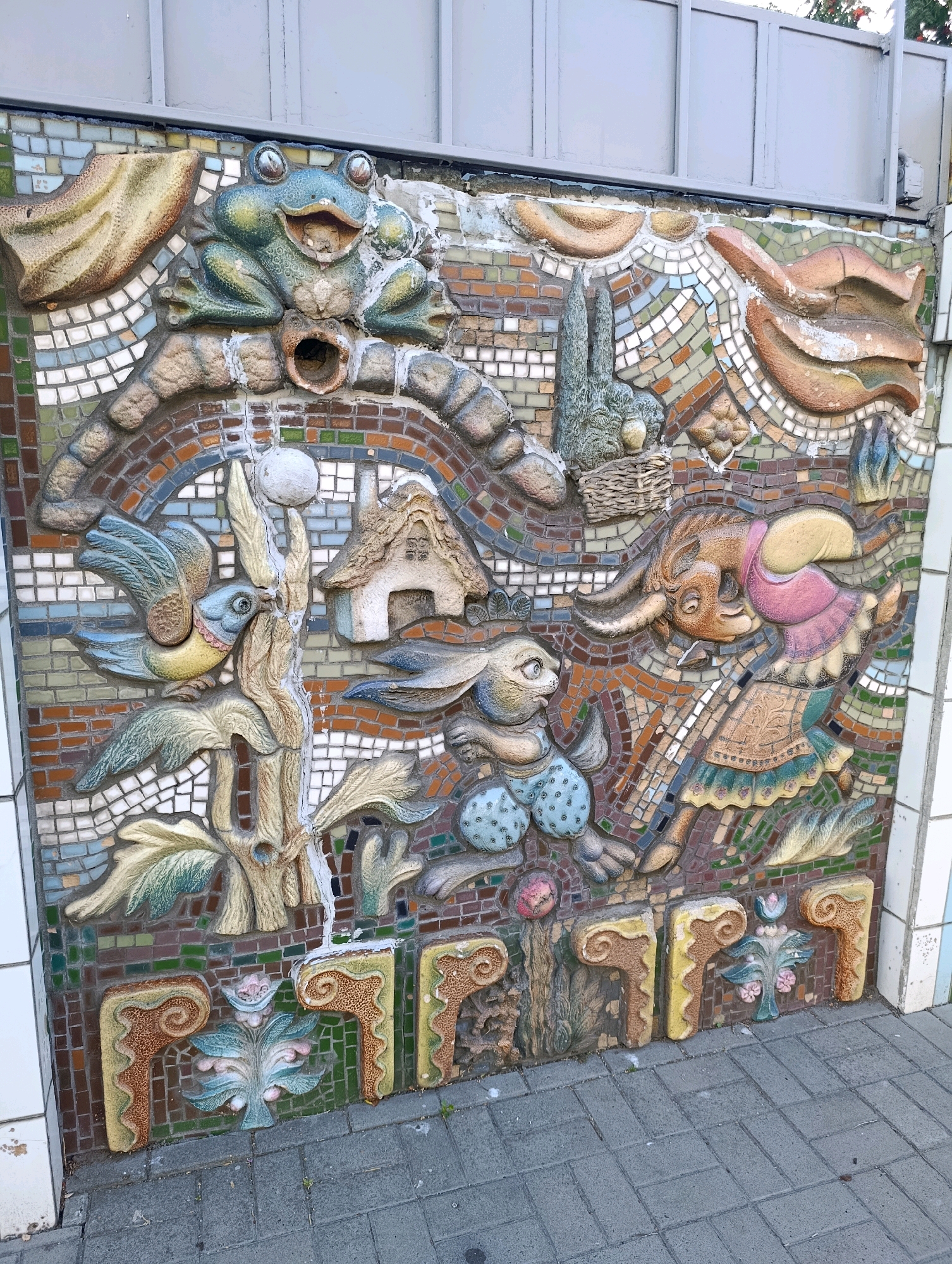 The image depicts a section of a colorful mosaic mural on an exterior wall. The mural features a whimsical scene with anthropomorphic animals and stylized natural elements. In the upper portion, a large green frog with wide eyes and an open mouth is prominent. Below the frog, a blue bird with outstretched wings is perched on a branch. To the right of the bird, a small house with a thatched roof is visible. The central figures include two rabbits, one in blue polka dot shorts and the other in a pink dress and green skirt. A brown creature with horns, also dressed in a pink garment, is depicted leaping. The lower section of the mural contains decorative floral motifs and architectural elements. The mural is constructed with a combination of mosaic tiles and three-dimensional sculpted figures. The wall is situated outdoors, with a grey tiled pavement in the foreground and a metal structure forming the upper border of the scene. This mural is located on the wall of the Cherkasy Regional Philharmonic Hall in Cherkasy, Ukraine.