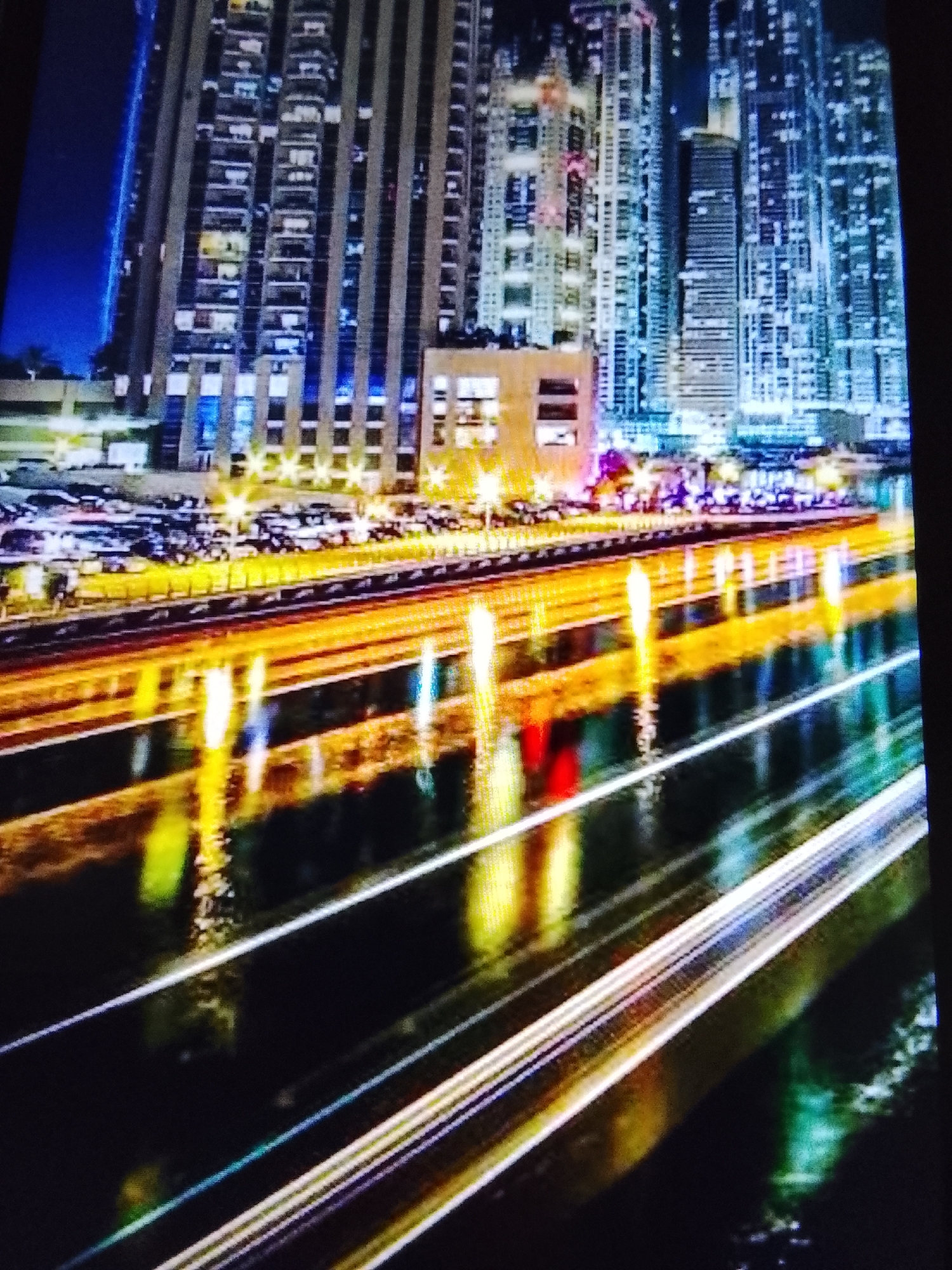 The image depicts a nighttime cityscape, characterized by a long exposure effect that blurs the lights of moving vehicles into streaks. Tall, modern buildings dominate the background, with many windows illuminated, suggesting a densely populated urban area. The foreground features a body of water, likely a river or canal, reflecting the city lights and the light trails from the vehicles, creating a vibrant, abstract display of color. The overall impression is one of a dynamic and lively city at night. The location is identified as Safonovo, Russia.
