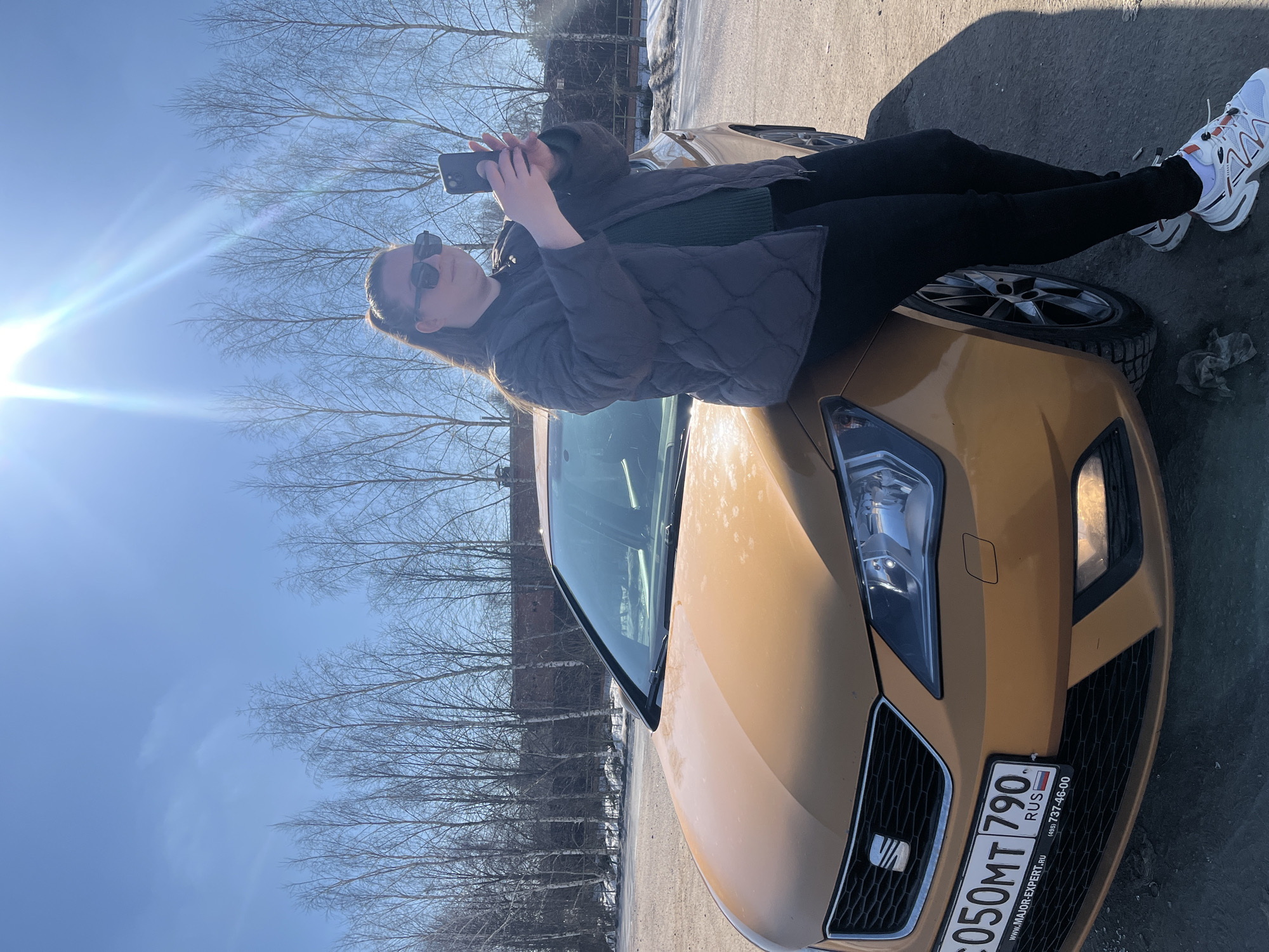 The image depicts an outdoor scene in Zubovo, Russia, under a clear, sunlit sky. A female subject is positioned to the right of a gold/orange two-door Seat Ibiza hatchback, leaning against its front driver-side fender. She has light brown hair pulled back in a ponytail, wears dark sunglasses, a dark gray quilted jacket over a dark green sweater, black pants, and white sneakers with orange and gray accents. The subject holds a black smartphone horizontally with both hands, pointed towards the left of the frame, indicating camera operation. The vehicle's front Russian license plate, "C050MT 790 RUS," is visible, identifying it as registered in the Moscow region. Its front grille, headlights, and a fog light are clearly seen. The ground beneath the vehicle and subject is a dark, unpaved surface, likely dirt or gravel, with scattered patches of snow or light-colored debris. In the background, a row of slender, bare deciduous trees lines the horizon under a vivid blue sky, with a prominent sun flare in the upper right quadrant. A dark, reddish-brown building with a chimney is partially visible behind the trees in the mid-ground.