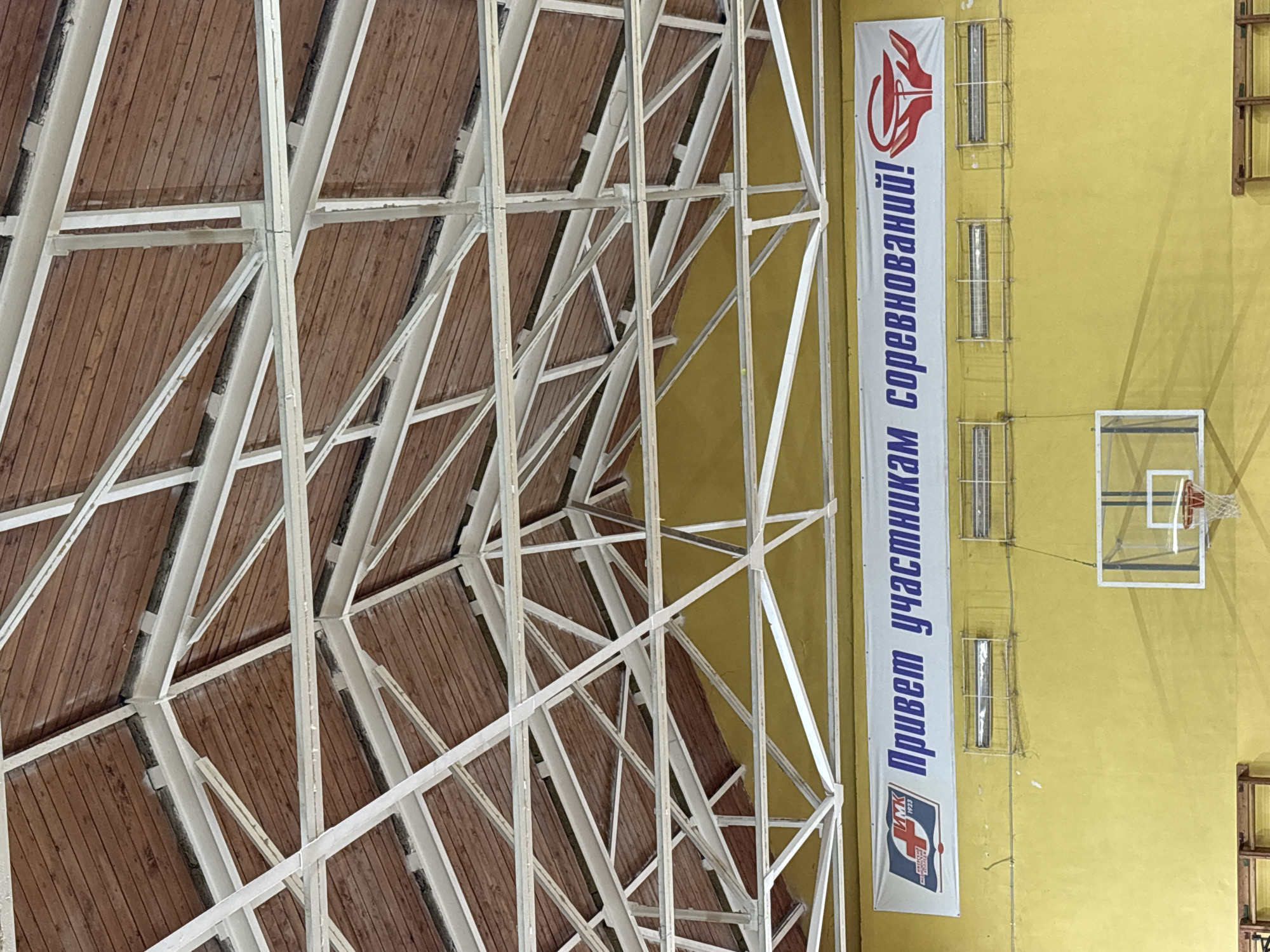 The image shows the interior of a sports hall, likely a basketball court, in Izhevsk, Russia. The camera is oriented towards the ceiling and a wall. The ceiling consists of a complex network of white metal beams supporting a wooden roof. The wall is yellow and features a vertical banner with Russian text and the logo of a sports organization, along with three sets of fluorescent tube lights housed in wire cages. A basketball hoop and backboard are mounted on the wall. A small wooden structure is present on the lower right of the wall.