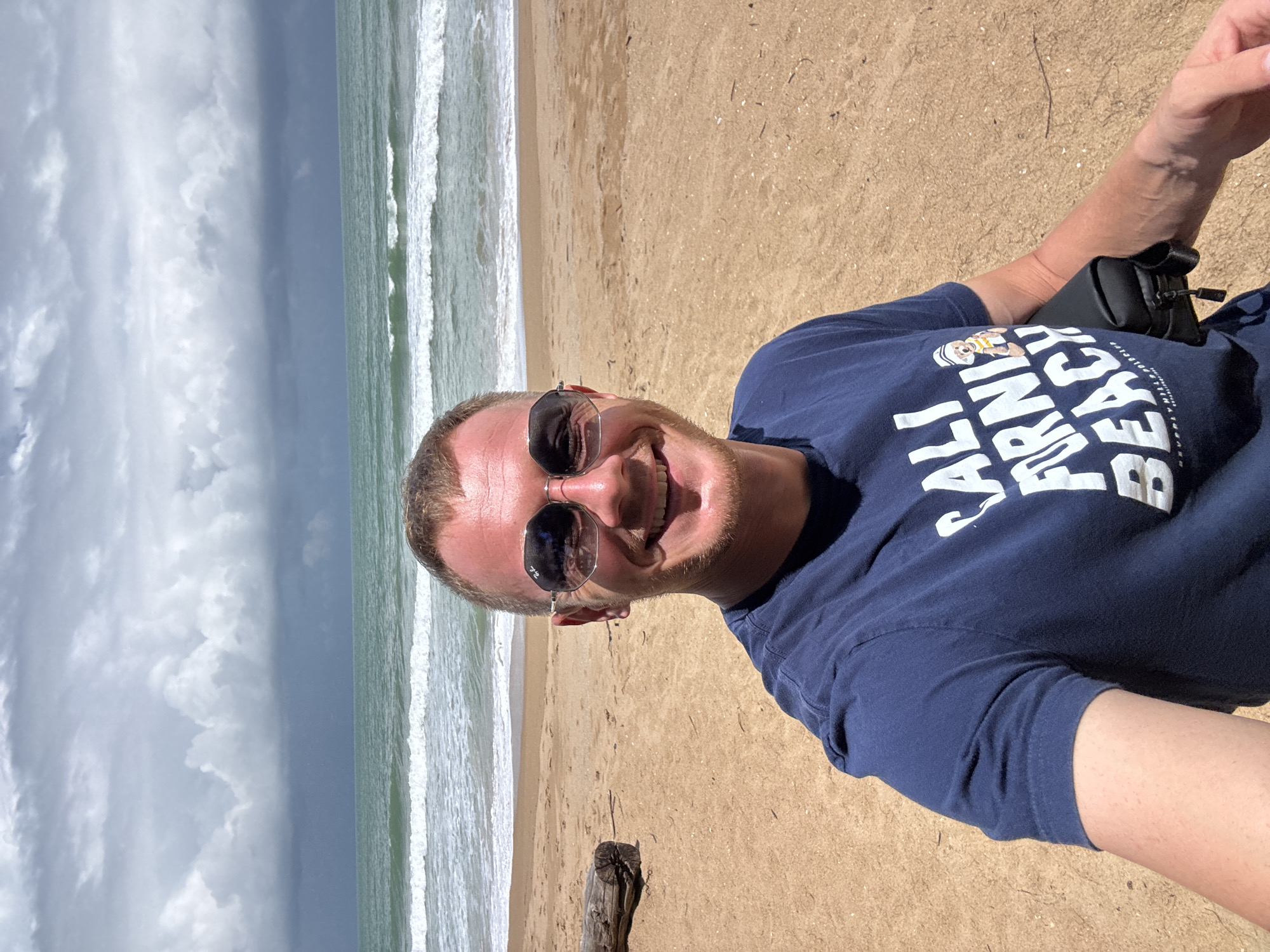A man is taking a selfie on a beach in Ban Khuk Khak, Thailand. The man is wearing a navy blue t-shirt with white text that reads "CALI FORNIA BEACH" and sunglasses. He has a light-skinned complexion and short, light brown hair. The beach sand is tan, and there is a piece of driftwood to the left of the man. The ocean is visible in the background with waves breaking on the shore. The sky is cloudy with dark clouds indicating possible rain.