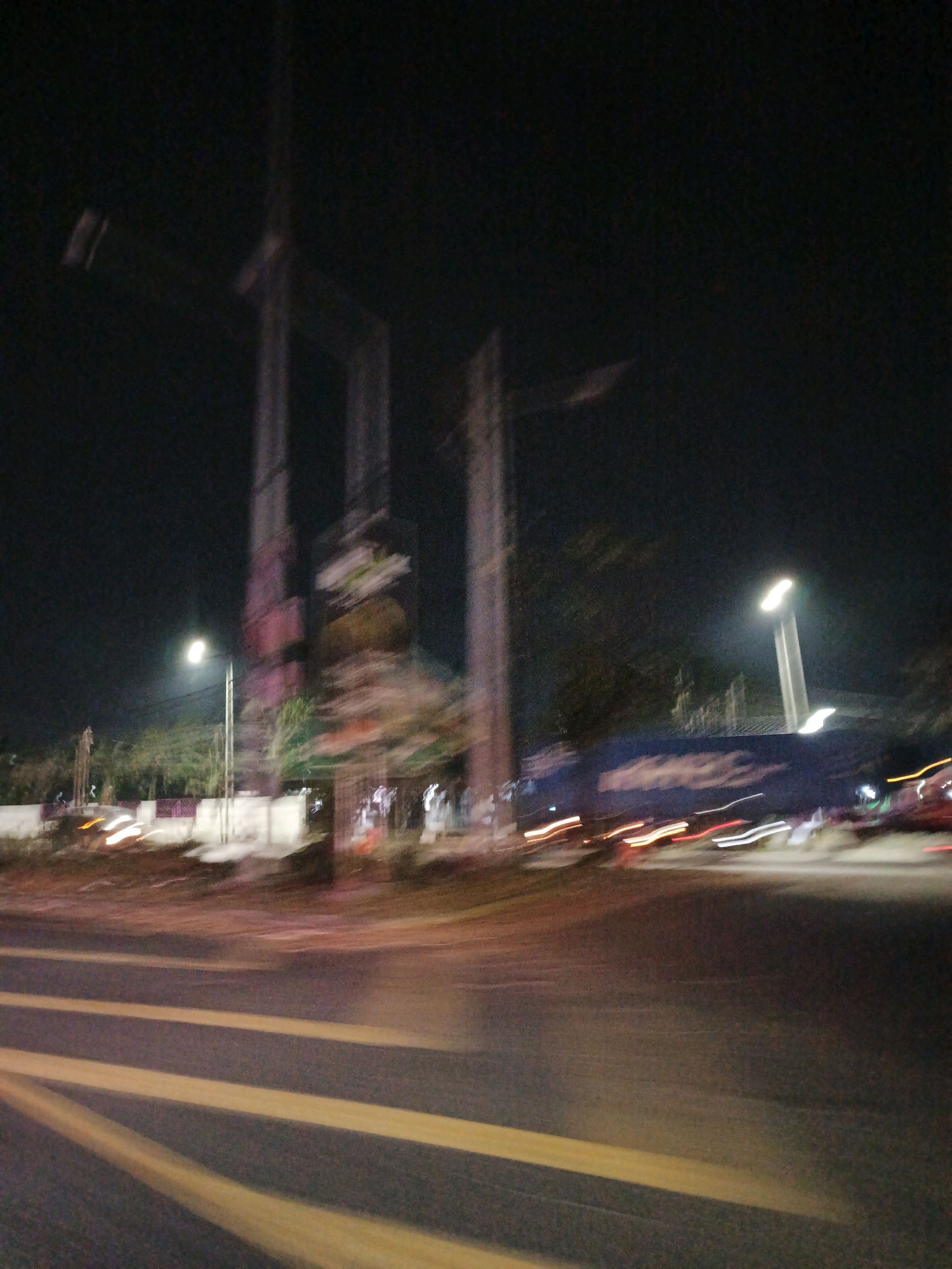 The image depicts a blurry night scene in Gempol, Indonesia. The foreground shows yellow road markings on dark asphalt, indicating lanes of traffic. In the midground, there are streaks of light from moving vehicles, suggesting a busy road. A large, dark truck with illuminated streaks of its lights is visible. To the left, a lamppost emits a bright light, and further back, the blurry shapes of trees and a white wall are discernible. In the background, the silhouette of tall structures, possibly construction cranes or buildings, is visible against the dark night sky. The overall impression is one of motion blur at night.