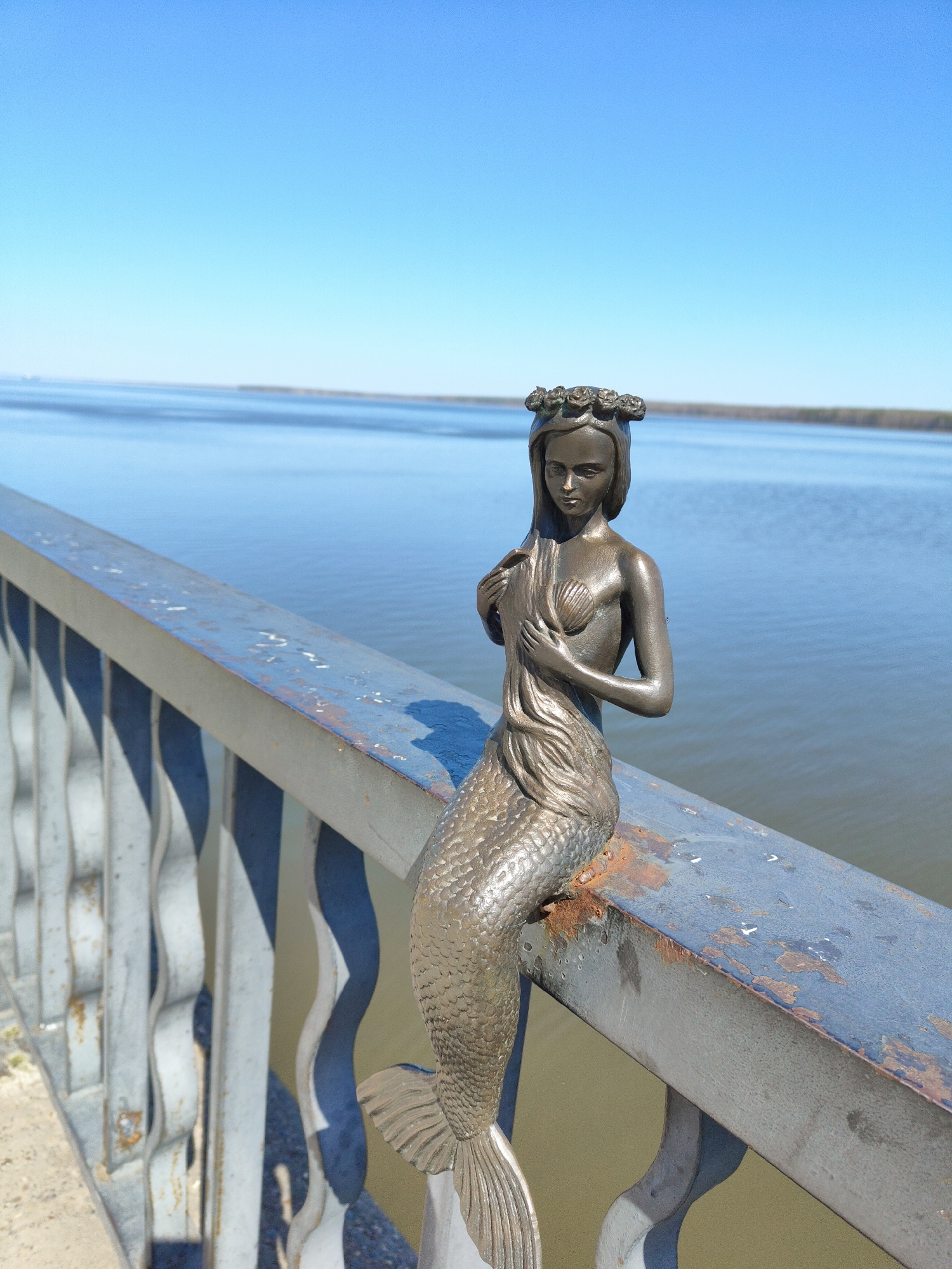 A bronze mermaid statue is perched on a blue metal railing in front of a large body of water under a clear blue sky. The railing appears to be part of a bridge or overlook. The mermaid has a detailed scale tail and a wreath of flowers on her head. Her arms are crossed in front of her, and she is gazing downwards. The metal railing is somewhat worn, with patches of rust. The body of water stretches to the horizon, where a strip of land can be seen. The location is identified as Unknown, Unknown.