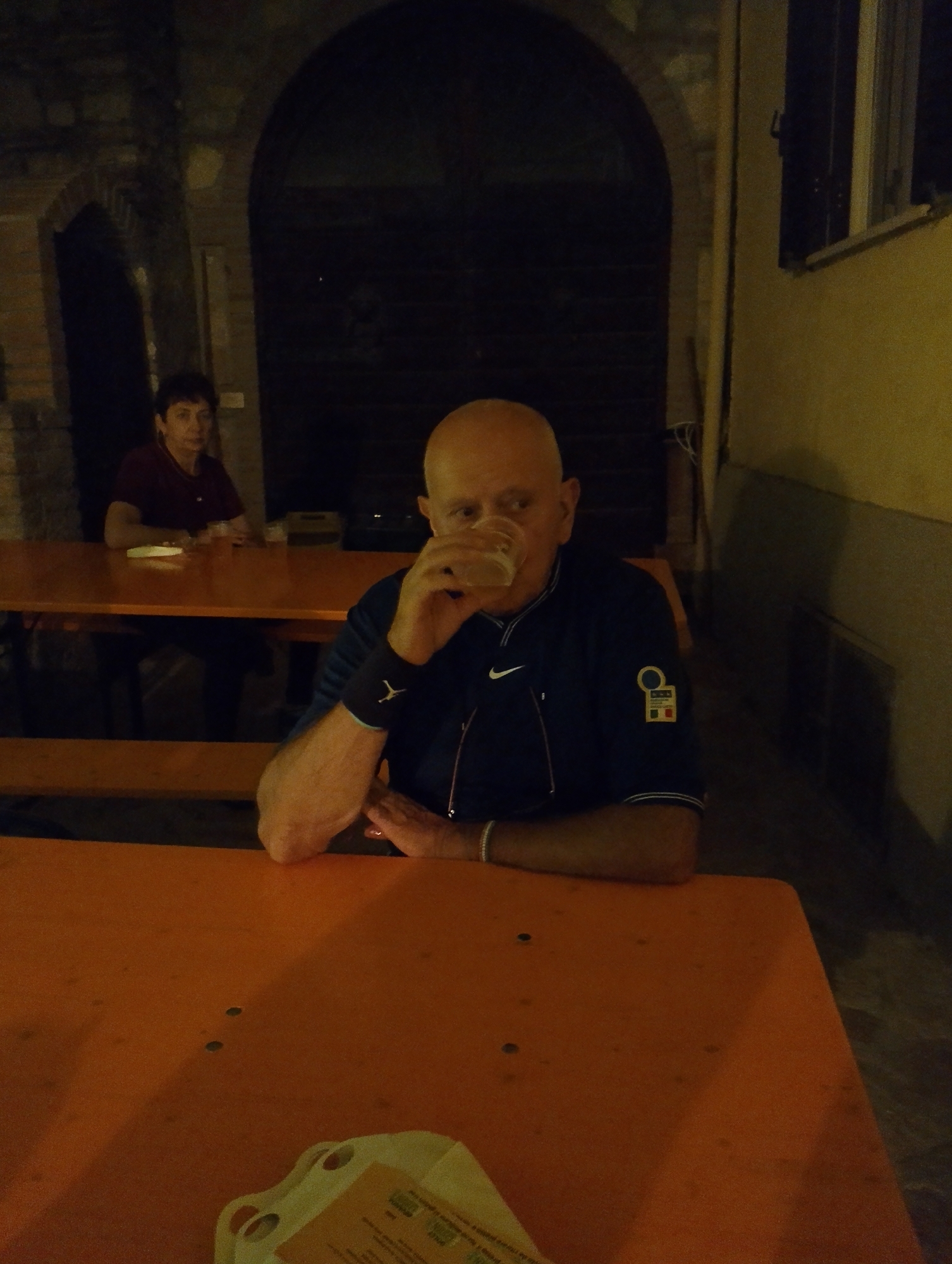 The image shows two people seated at an orange table outdoors at night. A bald man in the foreground is drinking from a glass of beer. He is wearing a dark blue shirt with a white Nike logo on the left chest and a circular emblem with Italian colors on the right sleeve. A woman with short brown hair is seated at a table in the background, wearing a dark red shirt. The setting appears to be a restaurant or bar with an arched entryway behind the woman. The location is identified as Rivodutri, Italy.