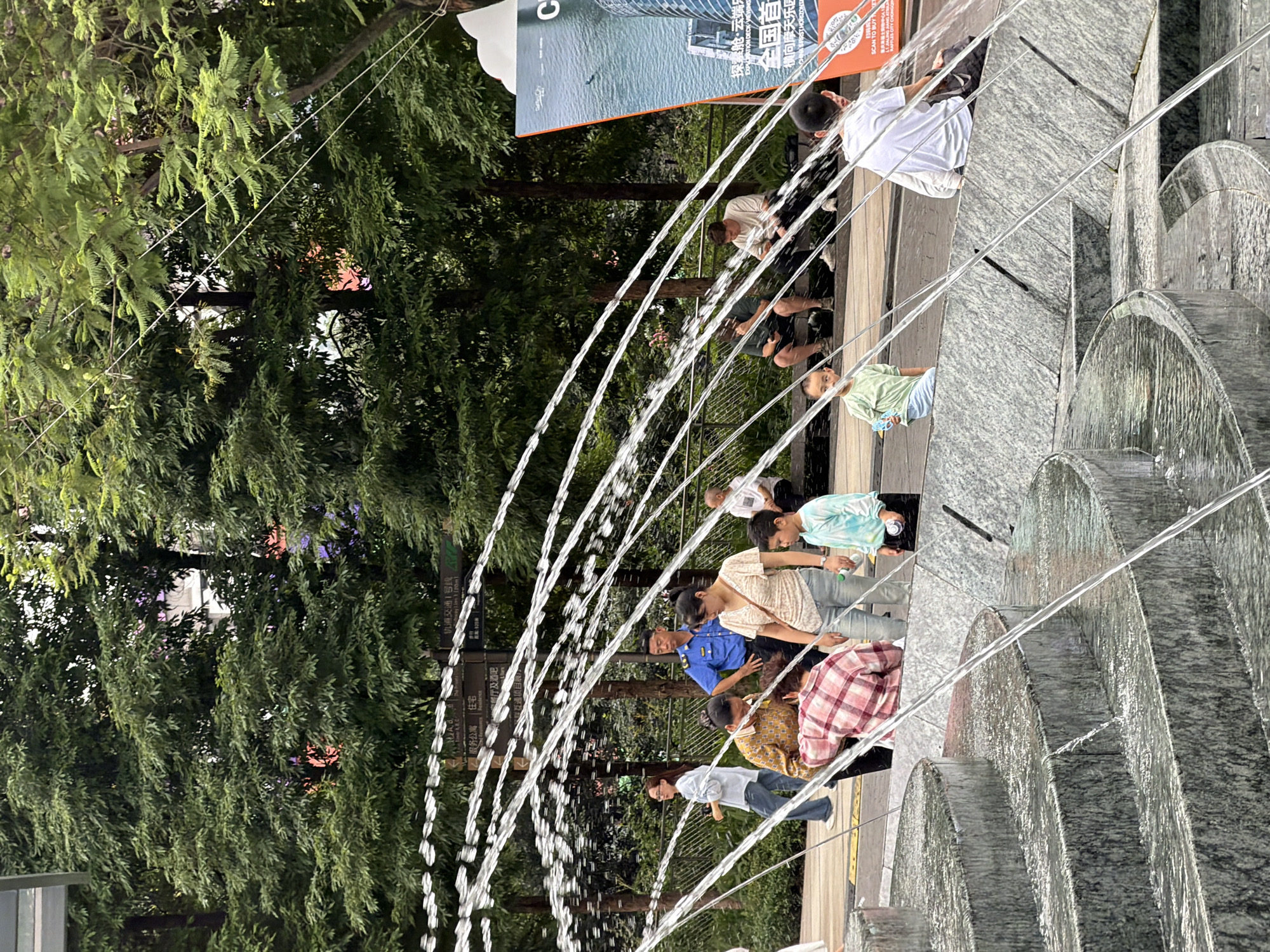 This image depicts a street scene in Yuzhong District, China, focusing on a public fountain with several people present. In the foreground, a tiered, stone fountain is active with water streams arching over the structures. Behind the fountain, a paved walkway is visible, bordered by lush green trees.
Several individuals are scattered throughout the scene. On the walkway, a woman in a white top and jeans walks to the left. Further back, a man in a blue uniform, possibly a security guard or police officer, stands looking forward. To his right, a woman in a light-colored top and jeans is walking. In front of them, a person wearing a red and white plaid shirt is bending over. A young boy in a light blue and green tie-dye shirt stands facing the camera, holding a small blue object.
Further back, several people are seated on a low wall or bench. Two men are visible in this group, one in a dark shirt and the other in a light t-shirt, both appearing to be looking at their phones. Another individual, a child in a light green shirt and blue shorts, is also seated. On the far right, a person in a white t-shirt is seated, facing away from the camera.
A large advertisement or poster is visible on the right side of the frame, featuring an image of a modern building and text in Chinese. The overall environment suggests a public urban space with recreational elements like the fountain and green areas, possibly a park or plaza.