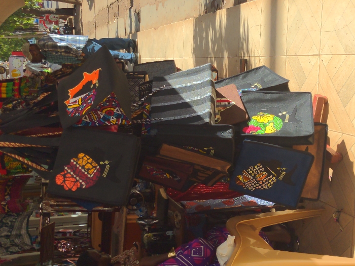 The scene is a street market in Lindi Street, Tanzania (latitude: -3.355, longitude: 37.341). A stack of colorful, handmade tote bags are displayed for sale. The bags feature intricate, patterned fabric designs. A woman wearing a colorful dress is seated in the foreground, while a man in a white shirt and brown pants stands in the background. The street is made of tiled squares, and there is a shadow from a nearby building on the sidewalk. The bags are resting on a wooden table with a brown surface.