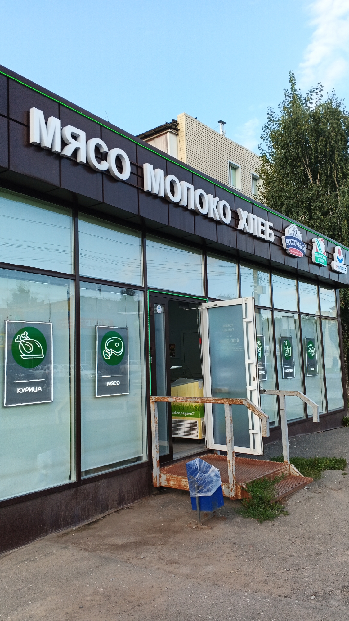 The image shows the exterior of a shop in Izhevsk, Russia, named "МЯСО МОЛОКО ХЛЕБ" (Meat Milk Bread). The storefront features large windows and white, three-dimensional lettering spelling out the shop's name. There are also smaller signs with green circular logos and text, one showing a chicken with the word "КУРИЦА" (Chicken) and another with a cut of meat and the word "МЯСО" (Meat). The shop has an open glass door, revealing a refrigerator unit inside with the text "свежее рядом" (fresh nearby). A rusty metal ramp with handrails leads up to the entrance. A blue trash bin with a plastic bag over it is positioned on the ramp. The sky is clear and blue, and a tree is visible to the right of the building. The overall scene depicts a typical street-level retail establishment.