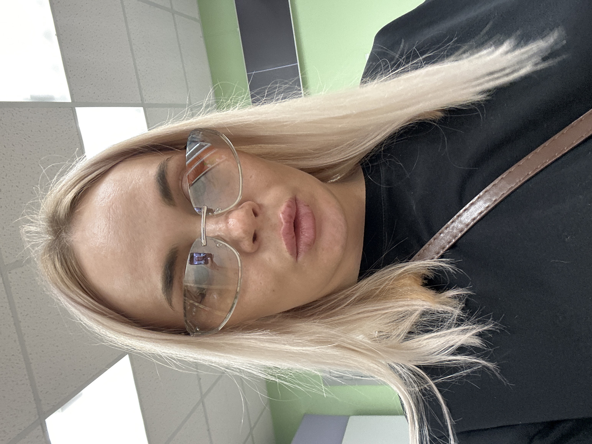 A woman with blonde hair and sunglasses is looking into the camera. She is wearing a black shirt. The background appears to be an indoor space with a tiled ceiling and light green walls. A brown strap is visible across her chest.