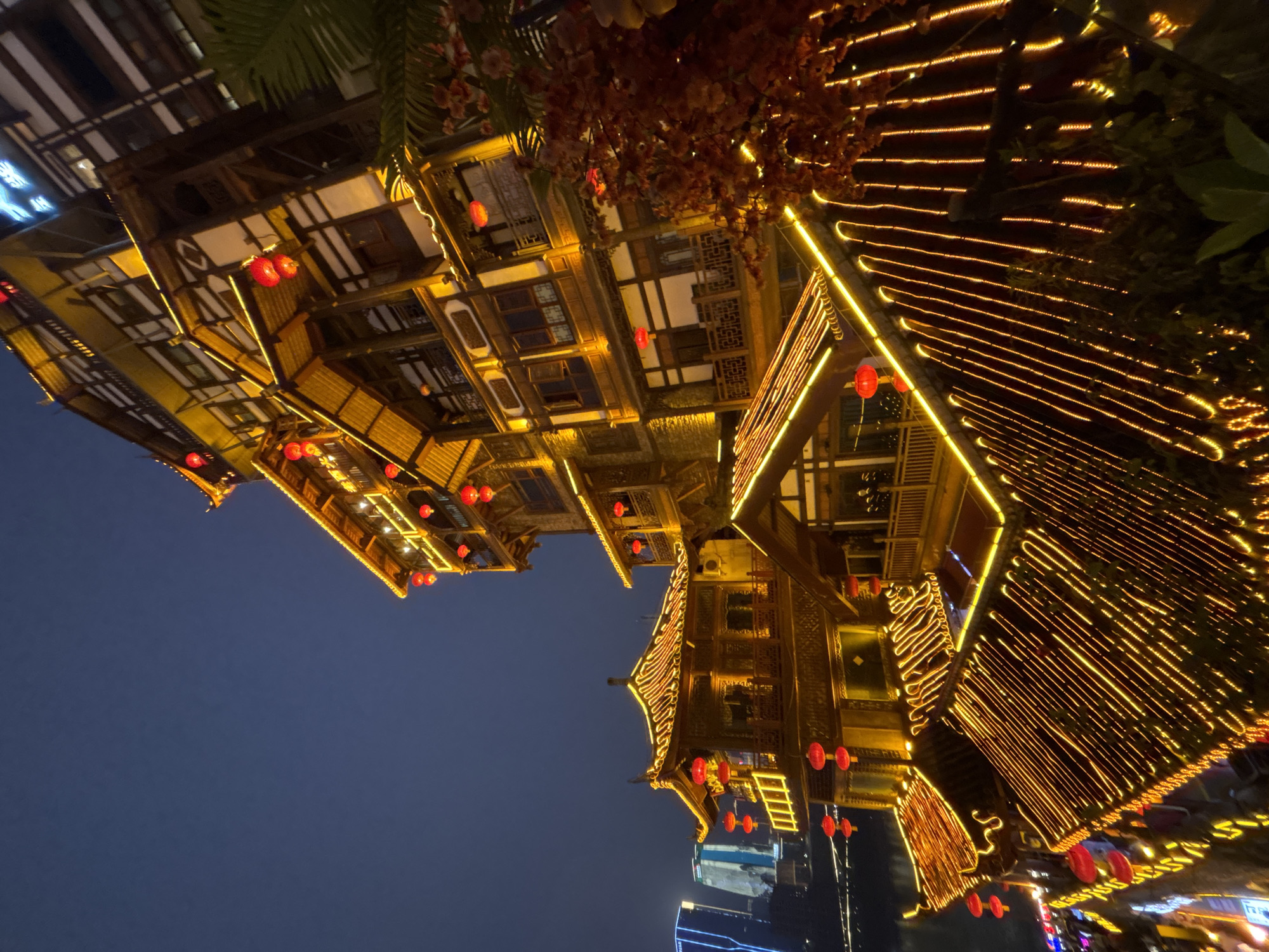 The image depicts a nighttime scene in the Yuzhong District of Chongqing, China, showcasing traditional Chinese architecture illuminated with bright lights. The buildings are adorned with strings of lights and red lanterns, creating a warm and festive atmosphere. The dark sky serves as a backdrop, emphasizing the vibrant glow of the buildings. Modern structures are visible in the distance, contrasting with the traditional architectural style. The image appears to be taken from a low angle, accentuating the height and grandeur of the buildings. The scene is an intersection of light, architecture, and culture within an urban setting.