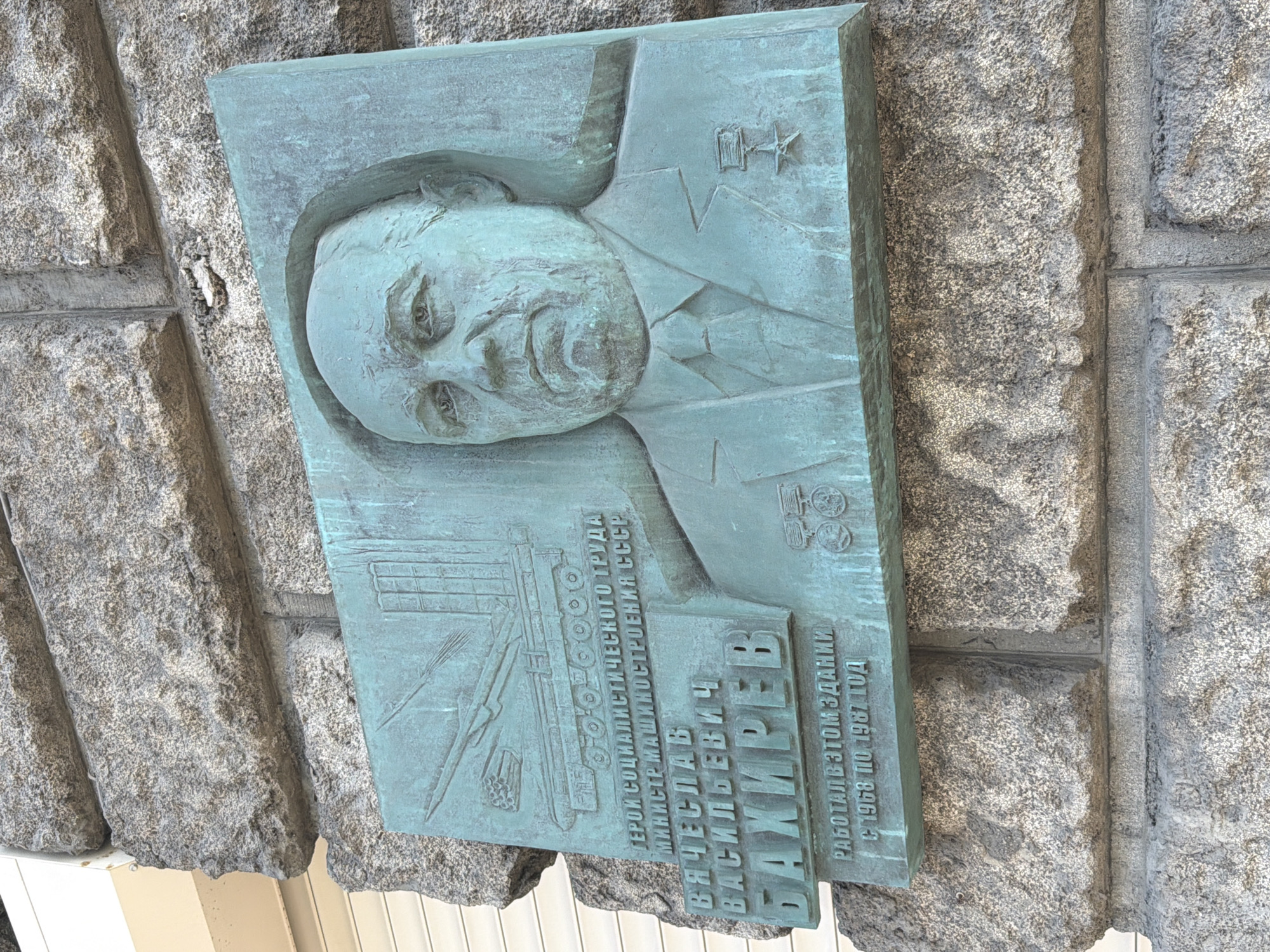 The image shows a bronze plaque affixed to a rough-hewn stone wall. The plaque depicts a bas-relief portrait of a man in a suit and tie. Above his head and to his left, are images of industrial equipment, and to the right are emblems of stars and other decorations. Below the portrait and images, text is inscribed in Cyrillic script. According to the text, the man depicted is Vyacheslav Vasilyevich Bakhirev, Hero of Socialist Labour, Minister of Mechanical Engineering of the USSR. The text also notes that he worked in the building from 1968 to 1987. The plaque is probably located in Moscow, Russia, where Bakhirev worked.