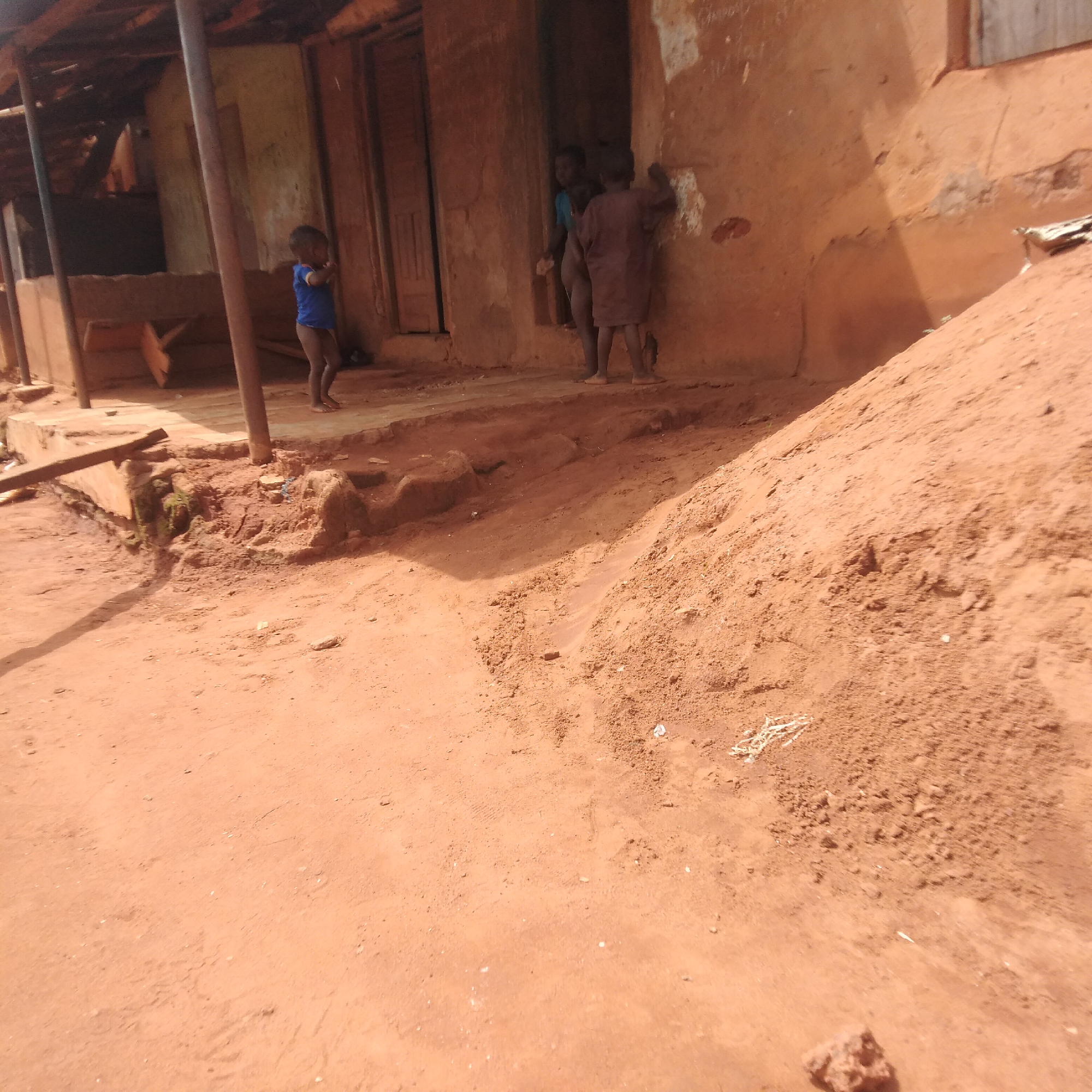 The image depicts a scene in an unspecified location within the city of Unknown, Unknown, with a traditional mud structure serving as the focal point. Three children are present. One child stands outside the building, while two others are near the entrance. The ground is predominantly reddish-brown soil. A large pile of earth or sand is visible in the foreground. The building appears to be constructed using traditional methods and materials.