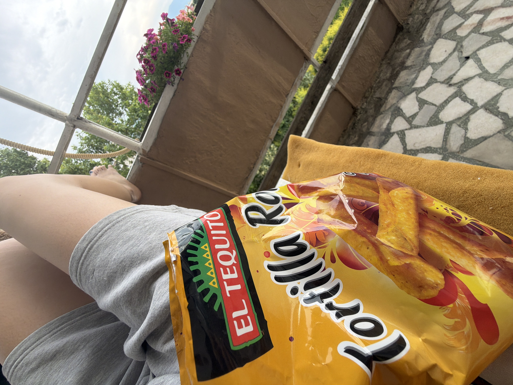 The image depicts a close-up view of a bag of "El Tequito Tortilla" chips held by a person. The person is wearing grey shorts and is seated. The background reveals an outdoor setting with a balcony or patio area. A railing, a planter with pink flowers, and trees are visible. The ground is covered with a mosaic of stones. The sky is cloudy. The scene appears to be a casual moment of relaxation. There is no specific information about the location in Pazardzhik, Bulgaria, provided in the image.