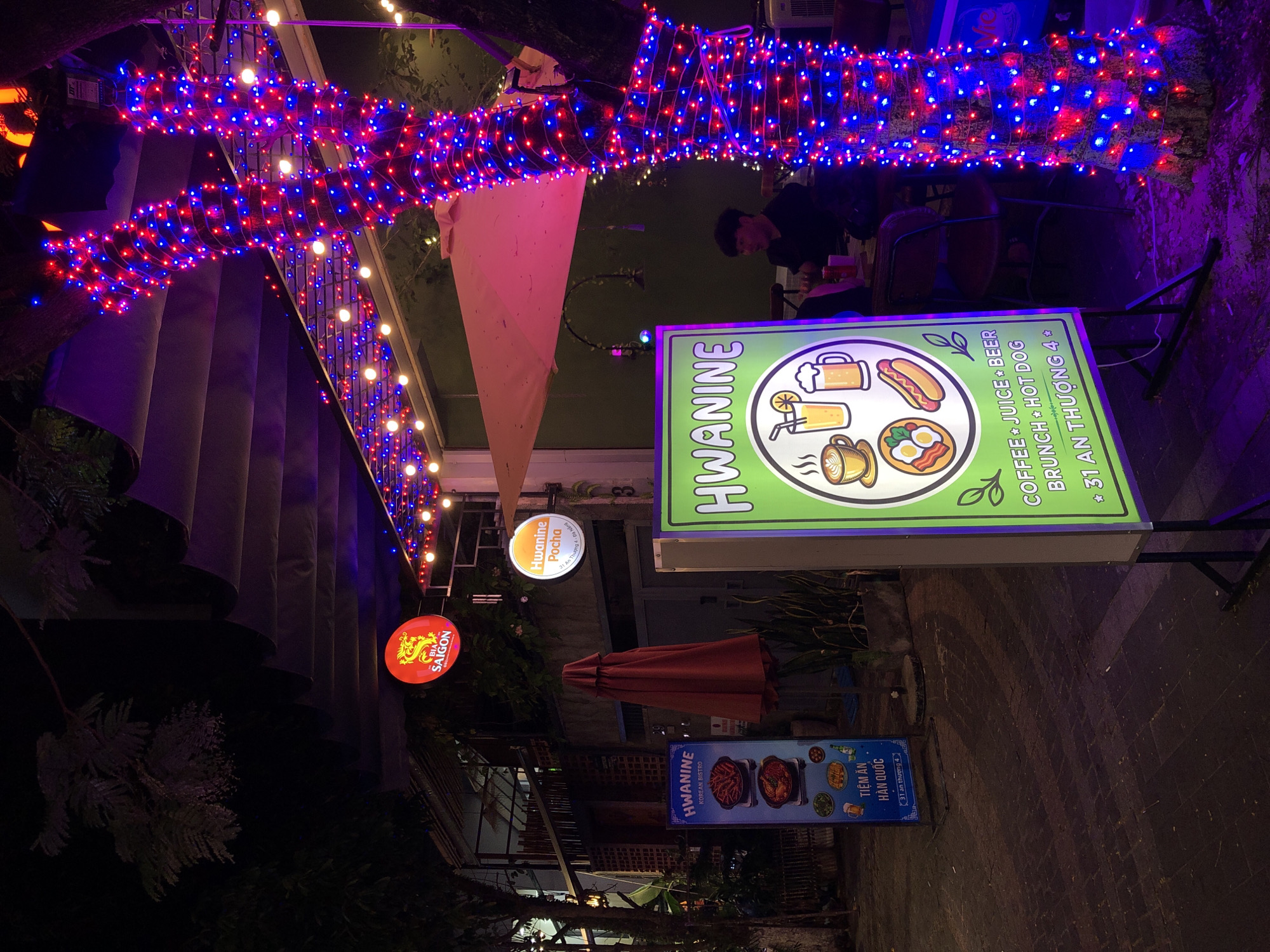 The image shows a street-level view of a restaurant or cafe at night in the city of Đà Nẵng, Vietnam. The establishment is decorated with numerous red and blue string lights wrapped around a tree and strung along an awning. A prominent sign for "Hwanine" with Korean text and images of food and drinks is visible in the foreground. To the left, another blue sign for "Hwanine Korean Bistro" with Vietnamese text is displayed. A red umbrella is partially deployed in the background. In the upper left, a circular red sign with "BIA SAIGON" is illuminated. A person is seated at a table in the background. The ground appears to be paved with cobblestones.