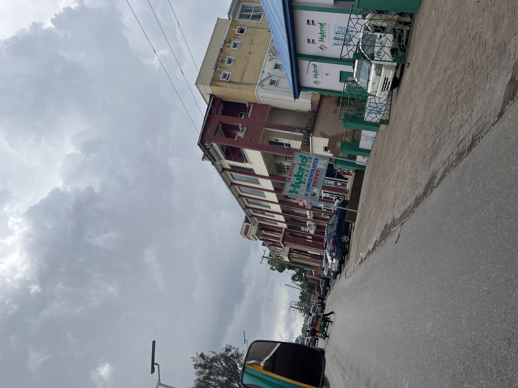 The media file depicts a street scene in Jos, Nigeria, under an overcast sky, suggesting a cloudy or rainy day. The perspective is from a vehicle, possibly a tuk-tuk or tricycle, looking forward and to the right.
Prominently featured are several buildings lining the street. One building has a yellow upper story with numerous small, square windows, and a red and white lower story with balconies and larger windows. Adjacent to this is a modern-looking building with green and white signage that reads "Hybrid FEEDS" and "Hybrid SEEDS." Another sign lower down on a building says "2x BETTER Hybrid FEEDS & CONCENTRATES DEPOT." A white van is parked in front of the building with the "Hybrid" signage.
The street is paved with asphalt and appears wet in places, consistent with recent rain. Several vehicles, including cars and a tuk-tuk, are visible on the road. People can be seen walking on the sidewalk and interacting with the vehicles. The overall mood is ordinary, with no strong emotional cues. The scene suggests a commercial or mixed-use area, likely during daylight hours given the brightness despite the cloud cover. The presence of utility poles and wires indicates an urban setting.