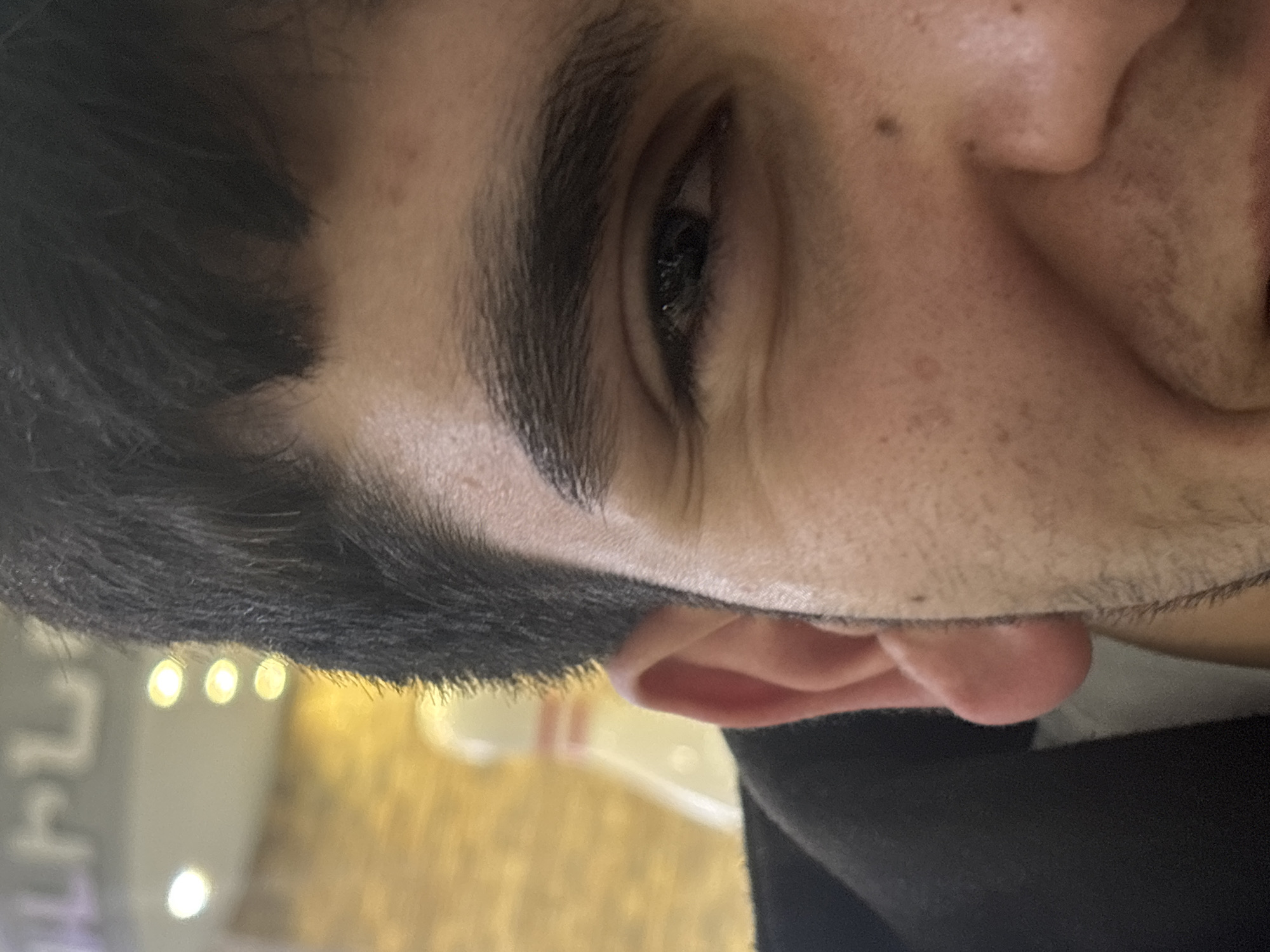 A close-up, Dutch-angled shot captures the left side of a person's face, likely male, in Yerevan, Armenia. The dominant features are a dark eye, prominent dark eyebrow, and a portion of the forehead, cheek, and ear. The person has dark hair and visible stubble or short facial hair along the cheekbone and jawline. The eye is open, looking slightly off-camera, with a neutral to contemplative expression.
The background is softly blurred with several circular, bright bokeh lights, indicating an indoor setting, possibly an evening or night scene in a public space like a restaurant, cafe, or shop. In the bottom left area of the background, there are indistinct, light-colored blurred shapes that could be stylized text or symbols, though they are not decipherable. No specific activity is discernible other than the person's presence in the location. The lighting is soft, accentuating the facial features against the blurry, illuminated background.