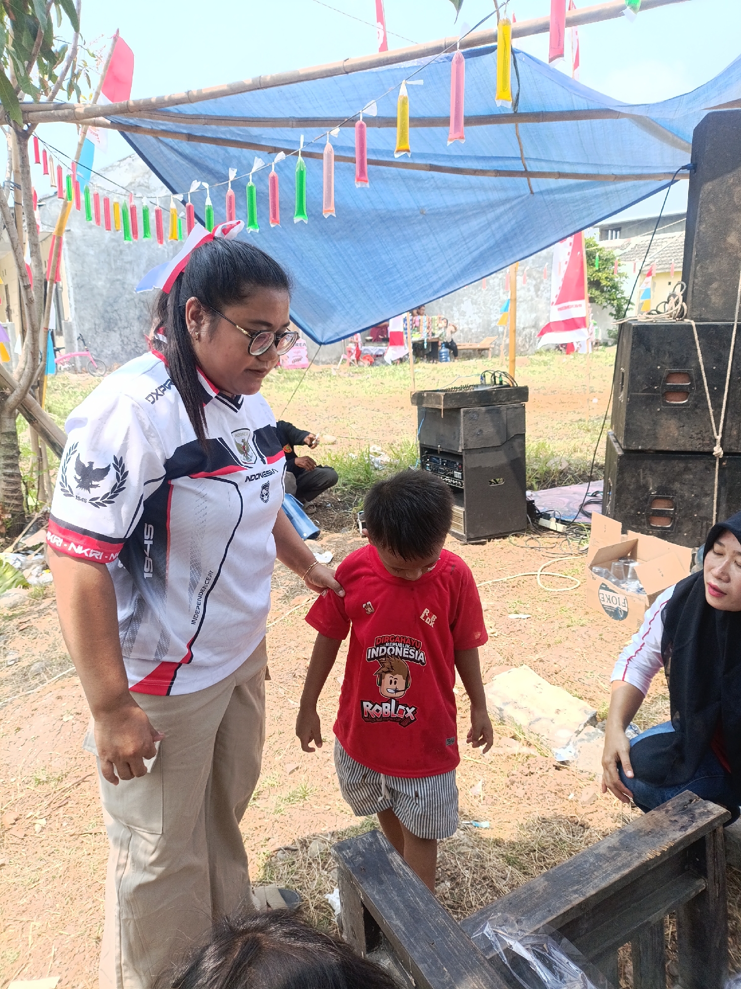 In Cariu, Indonesia, on a sunny day, an outdoor gathering is underway. A woman wearing a white and red athletic shirt with the Indonesian emblem and the year "1945" emblazoned on it, along with khaki pants, stands with her hand gently on the shoulder of a young boy. The boy is dressed in a red t-shirt featuring a Roblox graphic and "INDONESIA," paired with striped shorts. Behind them, a large blue tarp is strung between bamboo poles, decorated with colorful, water-filled plastic bags that resemble small popsicles. Red and white Indonesian flags are also visible, tied to poles and fluttering in the breeze.
The ground is covered with dry grass and dirt. To the right, stacks of large black speaker boxes are positioned, suggesting a sound system is set up. A mixing board sits on a platform near the speakers. A cardboard box containing plastic cups is also present. Further in the background, a few other individuals are visible, some sitting on wooden benches. A bicycle is parked near a building in the distance. The overall scene suggests a celebratory or community event in a rural or semi-rural setting.