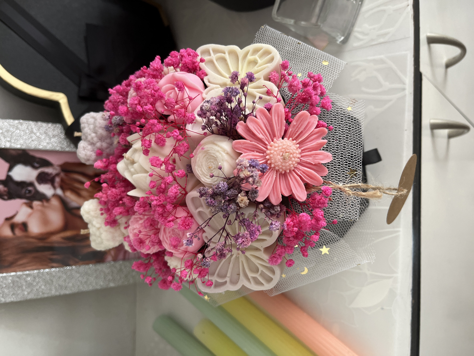 The image shows a close-up view of a decorative soap bouquet. The bouquet is arranged in a black cylindrical container, wrapped in grey mesh fabric with gold stars. The bouquet features a large pink gerbera daisy in the front, surrounded by white and pink soap flowers shaped like roses and butterflies. Smaller pink and purple dried flowers are interspersed throughout the arrangement. A kraft paper tag is tied to the bouquet with twine. In the background, a framed photograph of a woman kissing a dog is visible, along with several pastel-colored candles and a black box with a gold trim. The scene appears to be indoors, possibly on a shelf or table. There is no discernible indication of the location being in Topoli, Bulgaria.