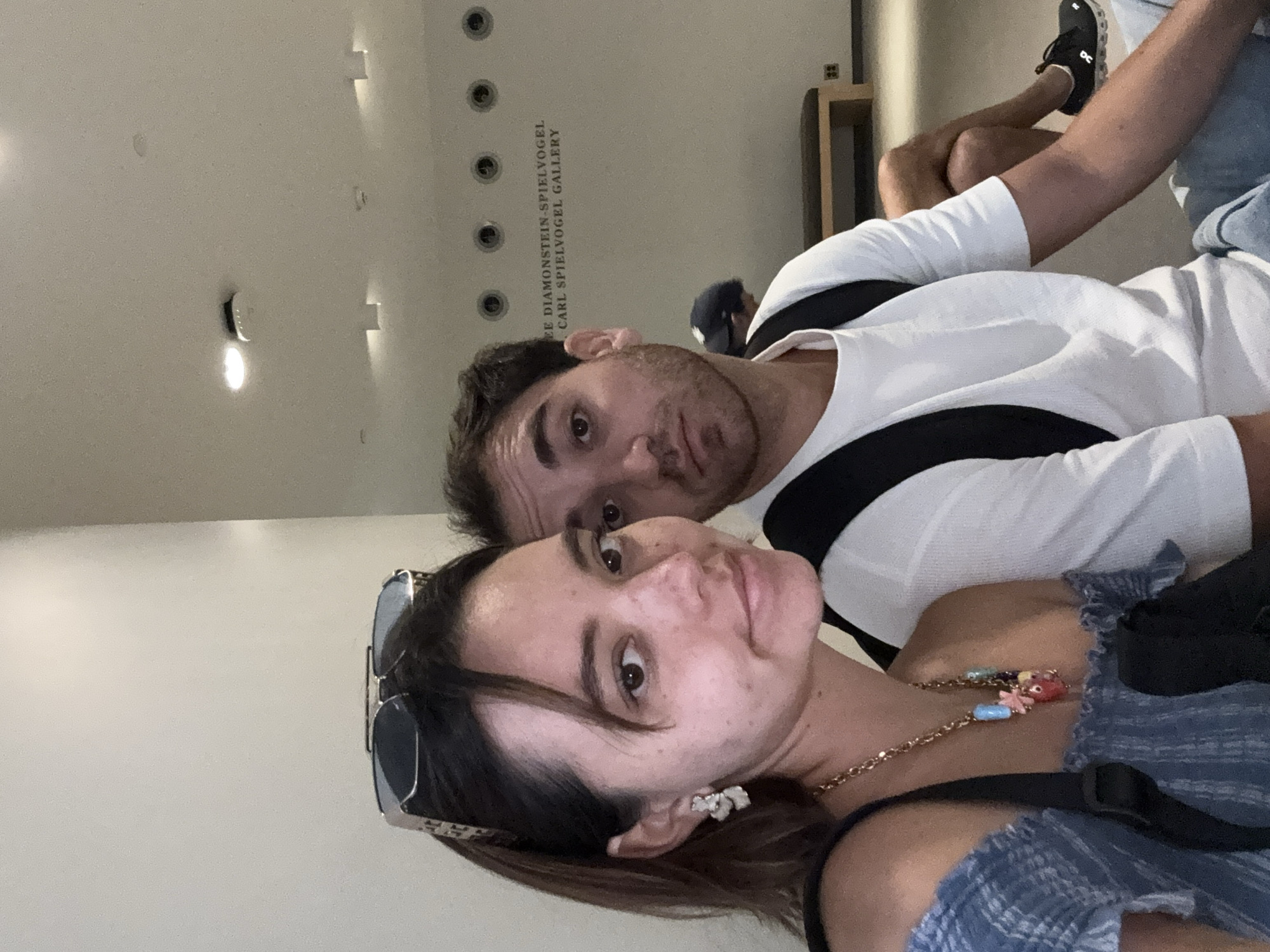 A man and a woman are posing for a selfie in what appears to be a museum or gallery, given the text on the wall behind them which reads "DIAMONSTEIN-SPIELVOGEL CARL SPIELVOGEL GALLERY". The woman is in the foreground, her face close to the camera, and she is wearing a blue off-the-shoulder top, a necklace with colorful charms, and large earrings. She has sunglasses perched on her head. The man is behind her and to her right, also looking towards the camera. He is wearing a white t-shirt and a backpack. In the background, there is a bench and circular lights on the wall. The overall lighting suggests an indoor setting. The location is identified as Washington, United States of America.