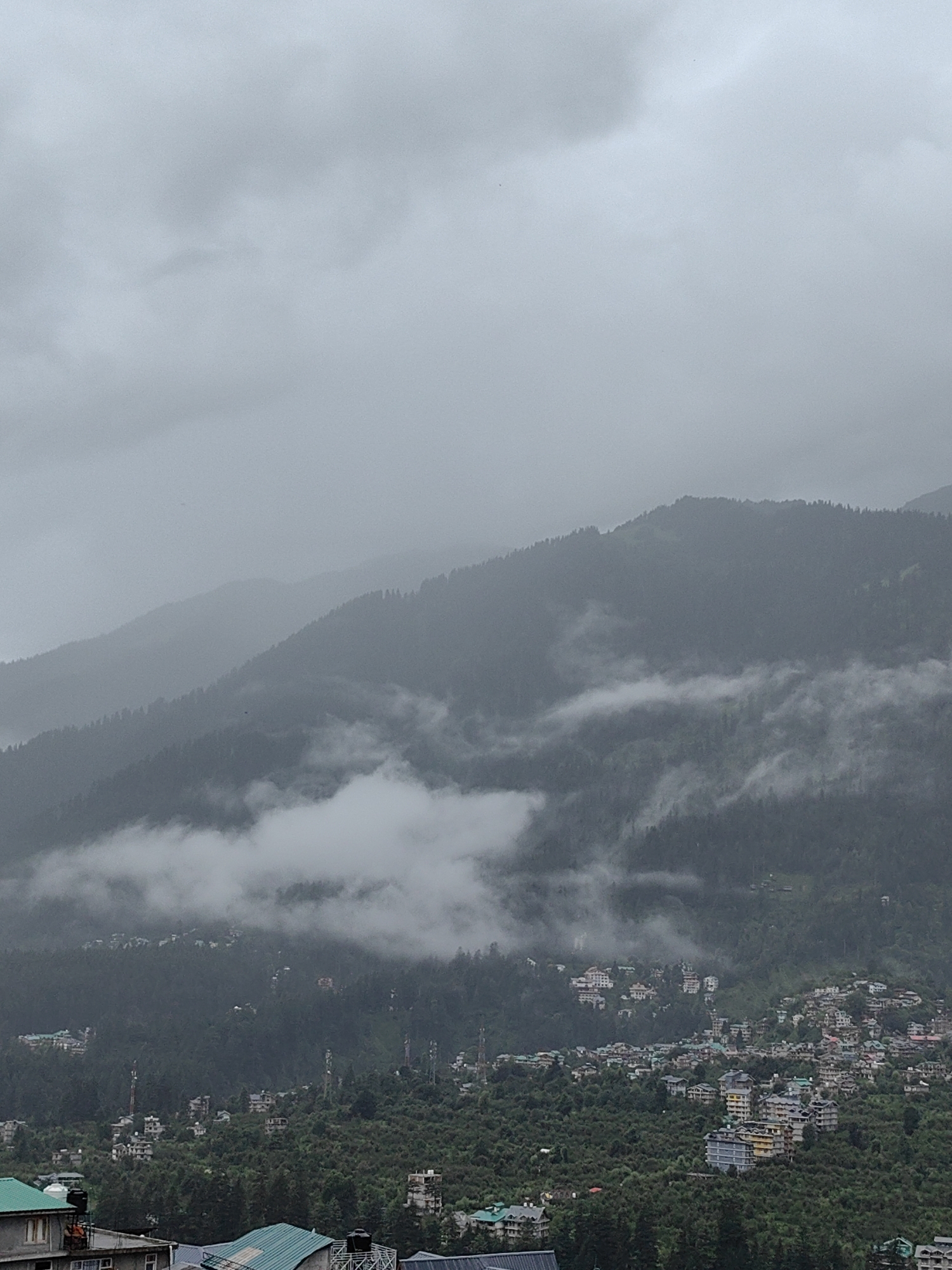 A high-angle, long shot captures a hazy, overcast scene of the town of Vashisht, India, nestled amongst lush, densely forested mountains. The sky is a uniform, muted gray, with wisps of fog or low clouds drifting through the mountain passes, obscuring the upper reaches of the peaks. A dense collection of buildings, ranging from multi-story structures to smaller dwellings, are visible spread across the verdant slopes, indicating a developed settlement. The architecture is varied, with many buildings featuring tiled roofs. Numerous trees, primarily dark green, cover the mountainsides and are interspersed with the buildings, creating a textured landscape. Several communication towers are discernible among the structures. In the foreground, the rooftops of a few more buildings are visible, some with distinctive green tiling and water tanks. The overall atmosphere is subdued due to the overcast weather. No people or specific actions are discernible in the image.