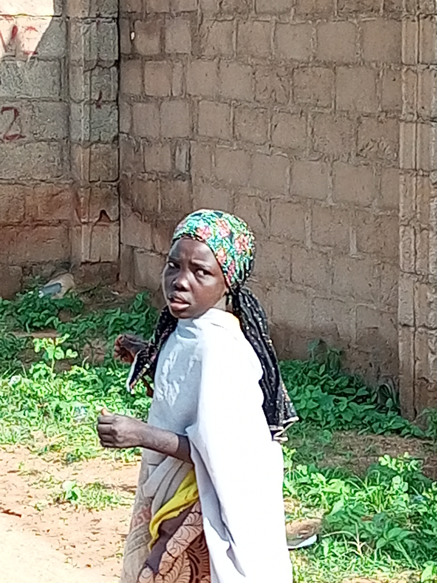 A young female, likely of Nigerian descent and possibly within the Tempure region, stands in front of a textured brick wall. She wears a vibrant floral headscarf, dark braids, and a light-colored tunic layered over a skirt with a patterned fabric. Her gaze is directed slightly to her left, and her mouth is slightly open. In her left hand, she holds a small, light-colored object. The background consists of lush green foliage and the rough texture of a brick wall, suggesting an outdoor environment.