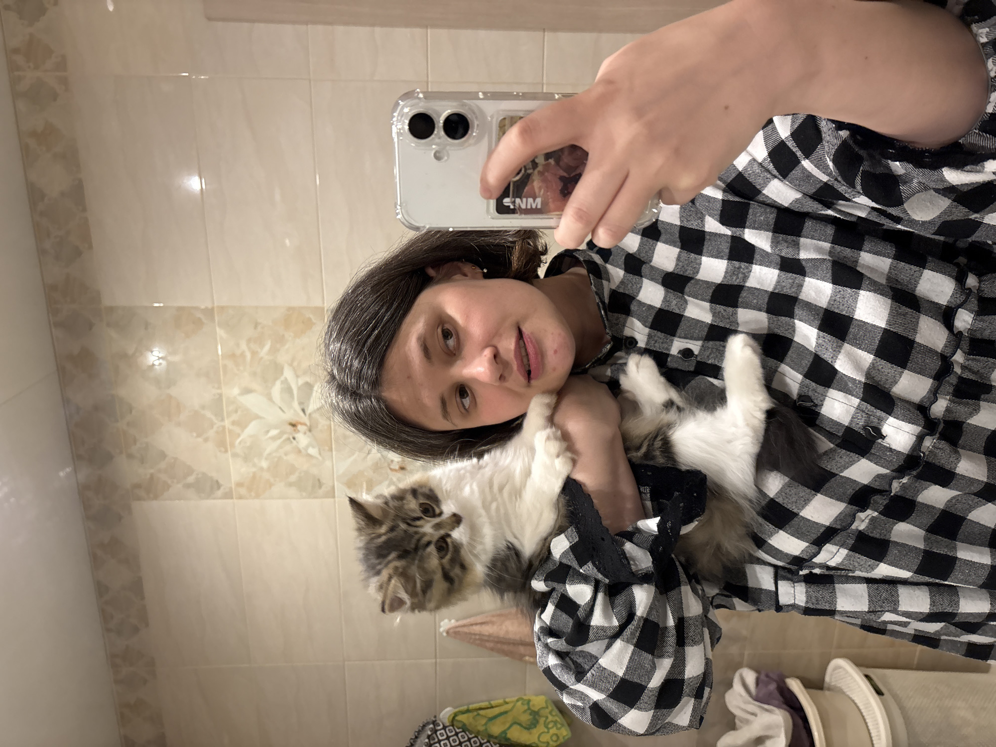 A woman is holding a cat while taking a selfie in a bathroom. The woman has short, dark hair and is wearing a black and white checkered shirt. The cat is white and gray. The woman is holding a white smartphone with a clear case. The bathroom has beige tiles with a decorative pattern. A portion of laundry is visible at the bottom. The location is in the city of Chaikovsky, Russia.