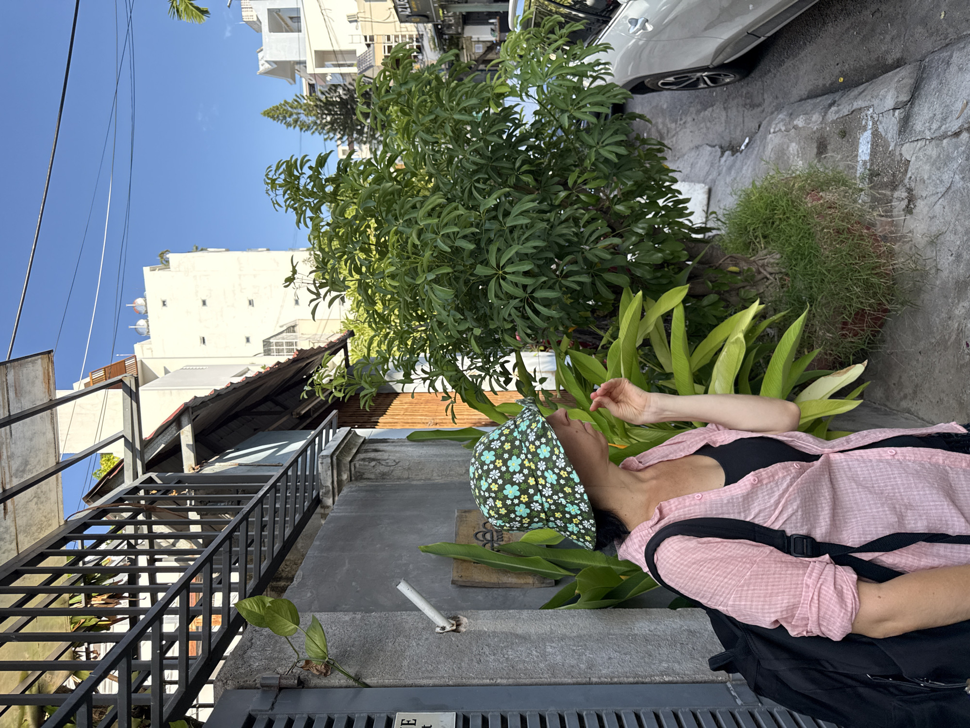 A person wearing a pink, short-sleeved button-down shirt over a black tank top and a patterned bucket hat stands on a sidewalk in Nha Trang, Vietnam. The person has a black backpack on their shoulders and is holding a finger to their lips. Behind them is a gray concrete wall with a black metal fence and a leafy green plant. A large green tree with many leaves is to the right of the person. In the background, there are white buildings and a clear blue sky. A white car is parked on the street to the right. The scene appears to be a street in a residential or commercial area.