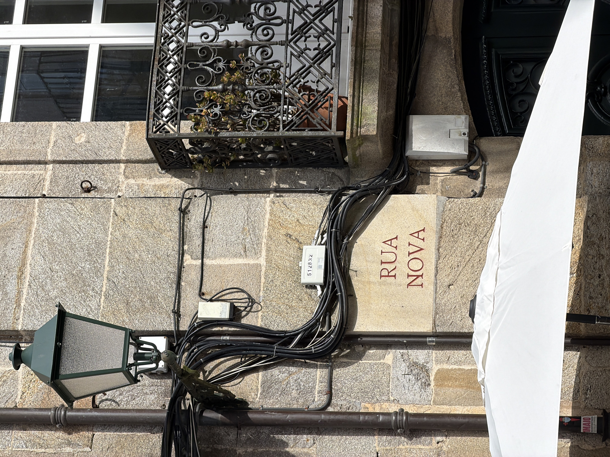 This is a photograph taken outdoors in Santiago de Compostela, Spain. The image focuses on a section of a stone building wall, featuring architectural elements and utility infrastructure. On the left, a dark green, ornate street lamp is mounted on a dark brown pipe. Below the lamp, a tangle of black and white electrical wires is bundled and routed along the stone facade. A small, white, rectangular utility box is also visible amongst the wires. Further to the right, a balcony with an intricate black wrought-iron railing encloses a potted plant. Above the balcony, a large white-framed window with multiple panes is present. A street sign with red lettering reads "RUA NOVA". In the foreground, the top portion of a large, white umbrella is visible, partially obscuring the lower part of the scene. The stonework of the building is rough and appears weathered.