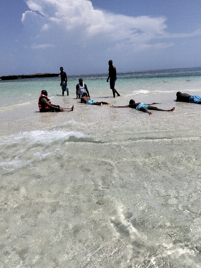The image depicts six adult males on a clear, shallow beach in Wasini, Kenya. The environment features transparent turquoise water revealing a light sandy bottom, with small waves breaking in the foreground. The sky is blue with a prominent white cloud formation in the upper left, and a dark, low-lying rocky landmass is visible on the distant left horizon.
Essential details of the individuals and their actions are as follows:
1. In the foreground-left, an individual wearing an orange life vest and dark shorts is seated on the submerged sand, partially immersed in the shallow water.
2. Standing slightly behind and to the right of the seated individual, another male holds a light-colored cloth draped over his arm.
3. A third male, wearing a white tank top and dark shorts, is crouched or kneeling in the shallow water in the mid-ground center.
4. Two individuals are lying prone (face down) in the shallow water. One is positioned in the mid-ground center, wearing blue and white patterned swim shorts, with arms extended forward.
5. The second prone individual is located further to the right in the mid-ground, also wearing blue and white patterned swim shorts.
6. A sixth male, wearing dark shorts, is walking towards the right in the mid-ground, with his back partially turned to the viewer.
No direct interactions between the individuals are observable; they appear to be individually situated or engaged with the water.