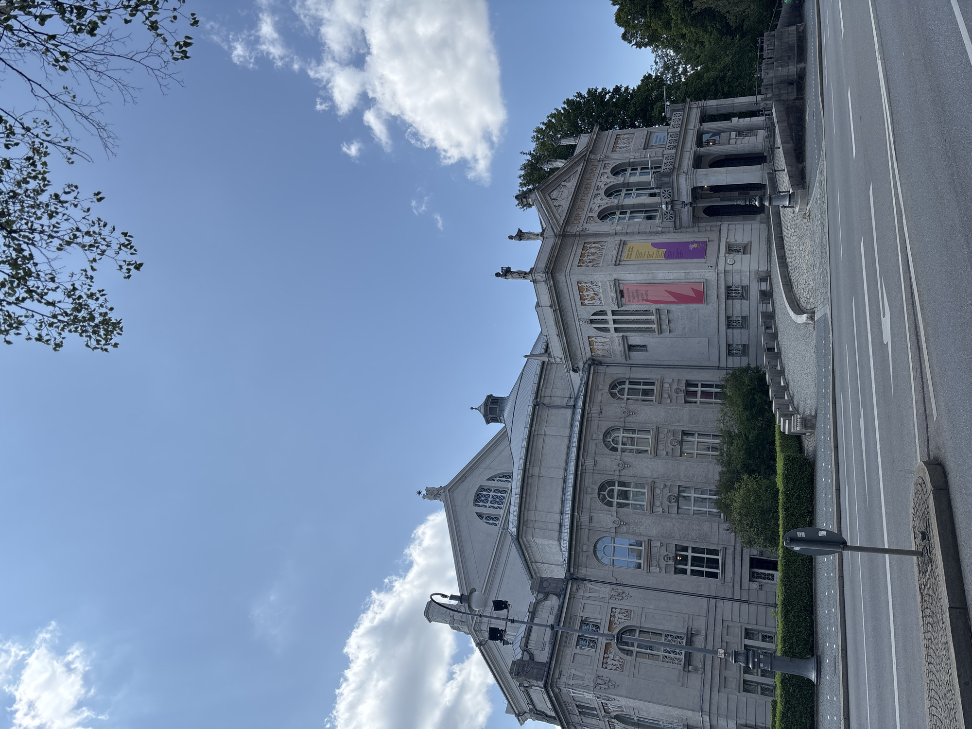 The image shows the exterior of a grand, historic building, likely a museum or opera house, located in Munich, Germany. The architecture is neoclassical, with a light gray stone facade, arched windows, and ornate decorative elements, including sculptures along the roofline. A prominent portico with columns leads to the entrance. Banners displaying text in German are hung on the facade. The building is situated on a street with a road and sidewalk. A lamppost stands on the sidewalk in front of the building. Trees and foliage are visible in the background, suggesting a park or green space. The sky is blue with scattered white clouds. The scene is captured from a street-level perspective, looking up at the building. There are no people visible in the immediate foreground, but the context suggests a public building in an urban setting. The overall impression is one of historical significance and architectural grandeur.