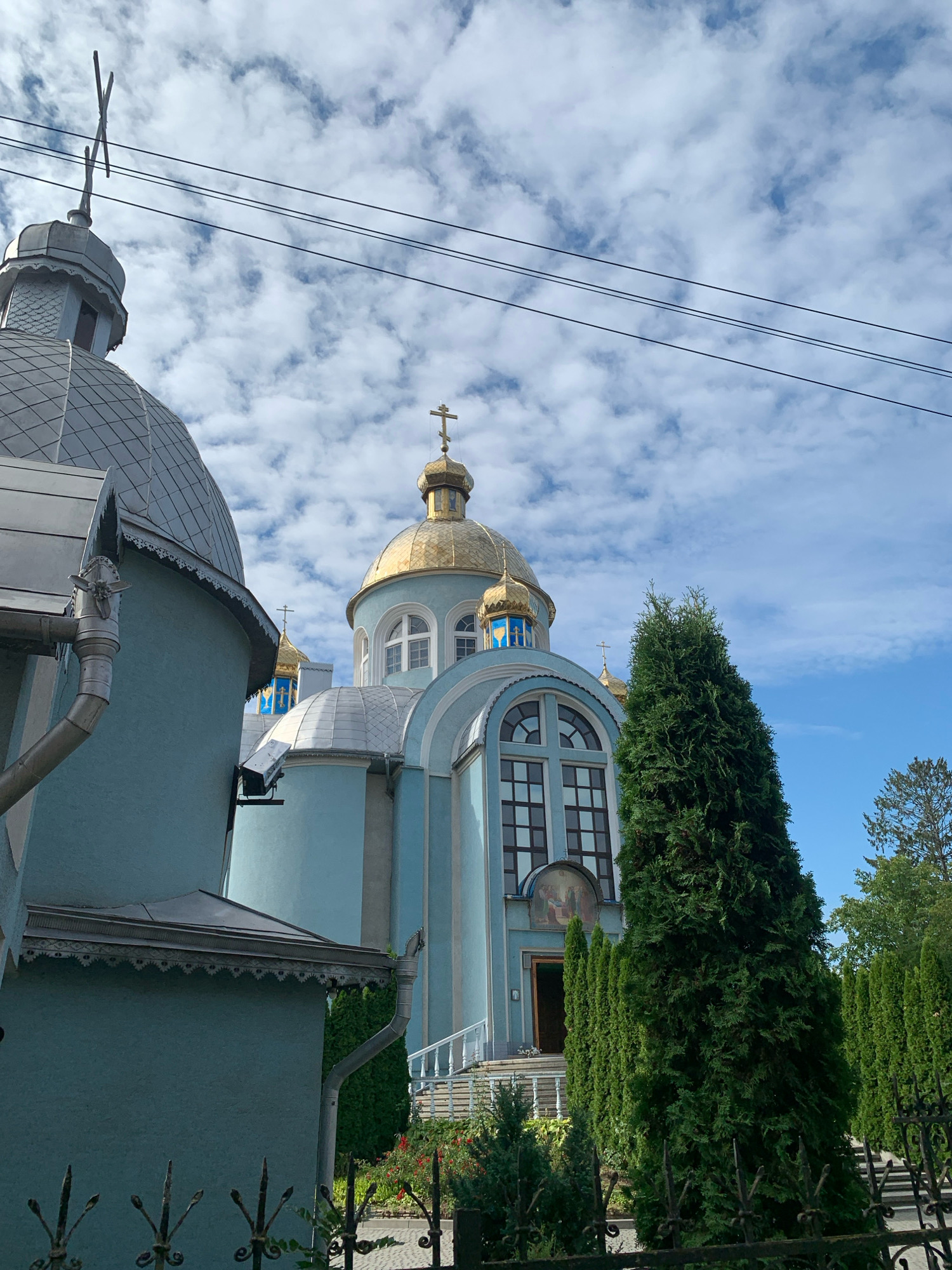 This image shows an exterior view of a light blue church with golden domes, located in Kolomyia, Ukraine. The church features multiple domes, including a prominent central dome topped with a cross. Arched windows with multiple panes are visible on the church's facade, and an open doorway leads into the building. The church is surrounded by lush green trees, some of which are tall and conical in shape, and a black wrought-iron fence runs along the foreground. The sky above is a vibrant blue, dotted with white, fluffy clouds. The perspective suggests the photo was taken from a street-level viewpoint, looking up towards the church structure.