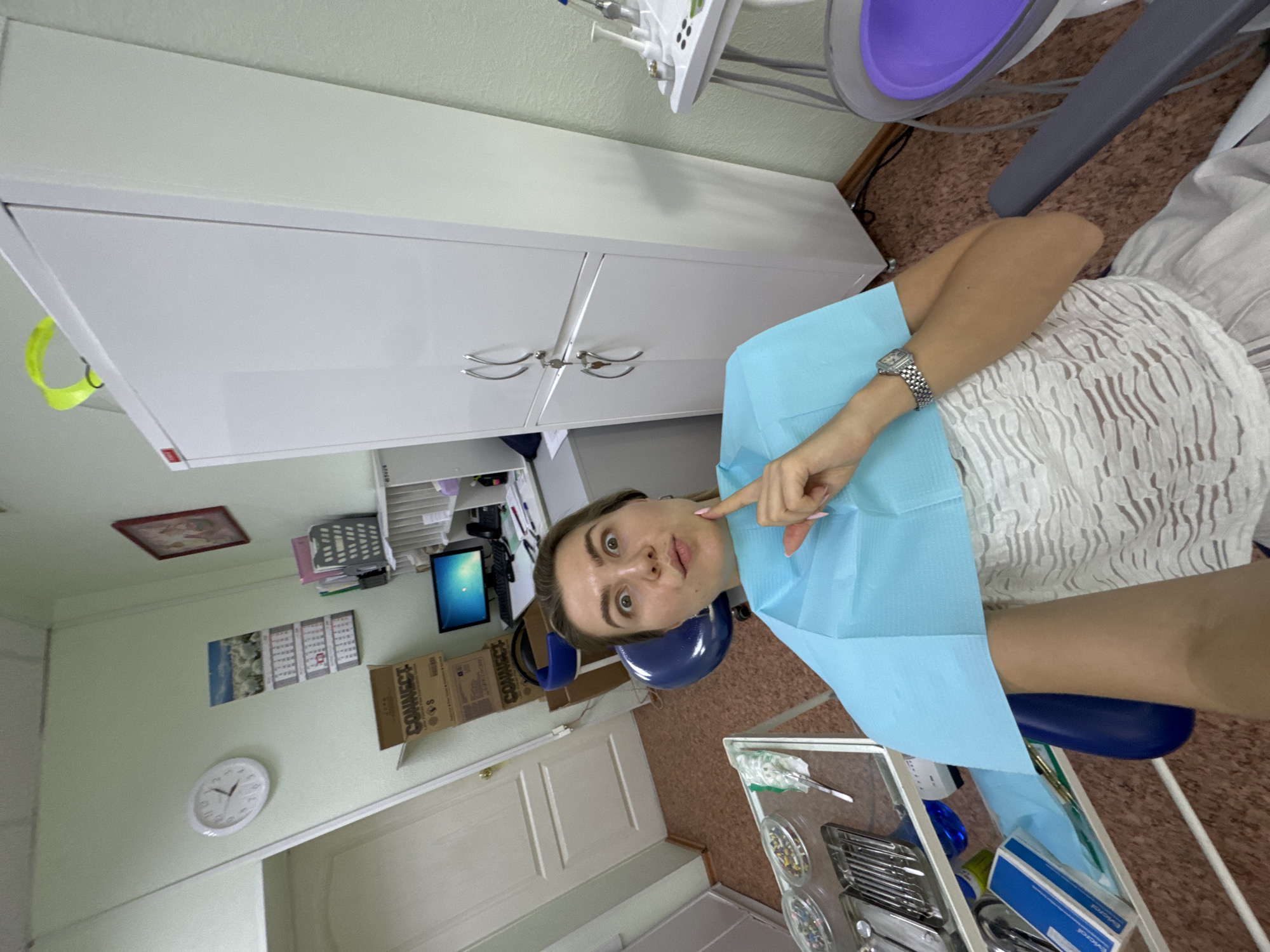 A woman with brown hair and light skin is seated in a dentist's chair, wearing a blue bib and a white patterned top. She is looking at the camera with a surprised expression, holding a finger to her chin. The room appears to be a dental office, with a white cabinet to the right, a clock on the wall, a computer monitor on a desk, and medical instruments on a tray in the foreground.