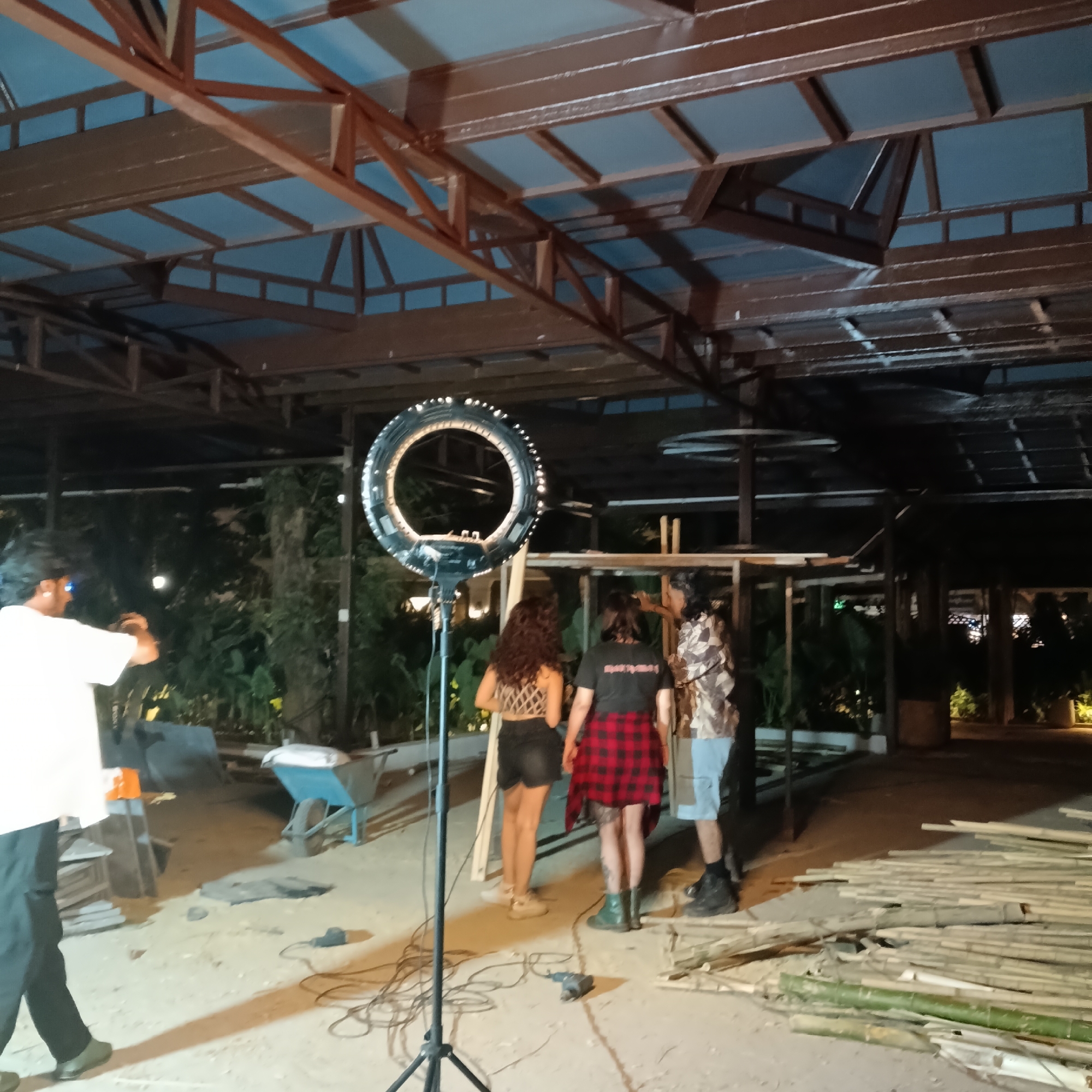 A ring light on a stand is in the foreground, illuminating three individuals who are standing in a semi-outdoor location. The first person on the left is wearing a black and white patterned top with black shorts. The second person is wearing a black shirt, a red and black plaid shirt tied around their waist, and green boots. The third person is wearing a camouflage shirt and blue shorts. A man in a white shirt and black pants is standing to the left and appears to be taking photos. There is construction occurring; some materials are scattered on the ground to the right. There is a partial structure behind the three individuals and a metal roof over the area. The image was taken in Panaji, India.