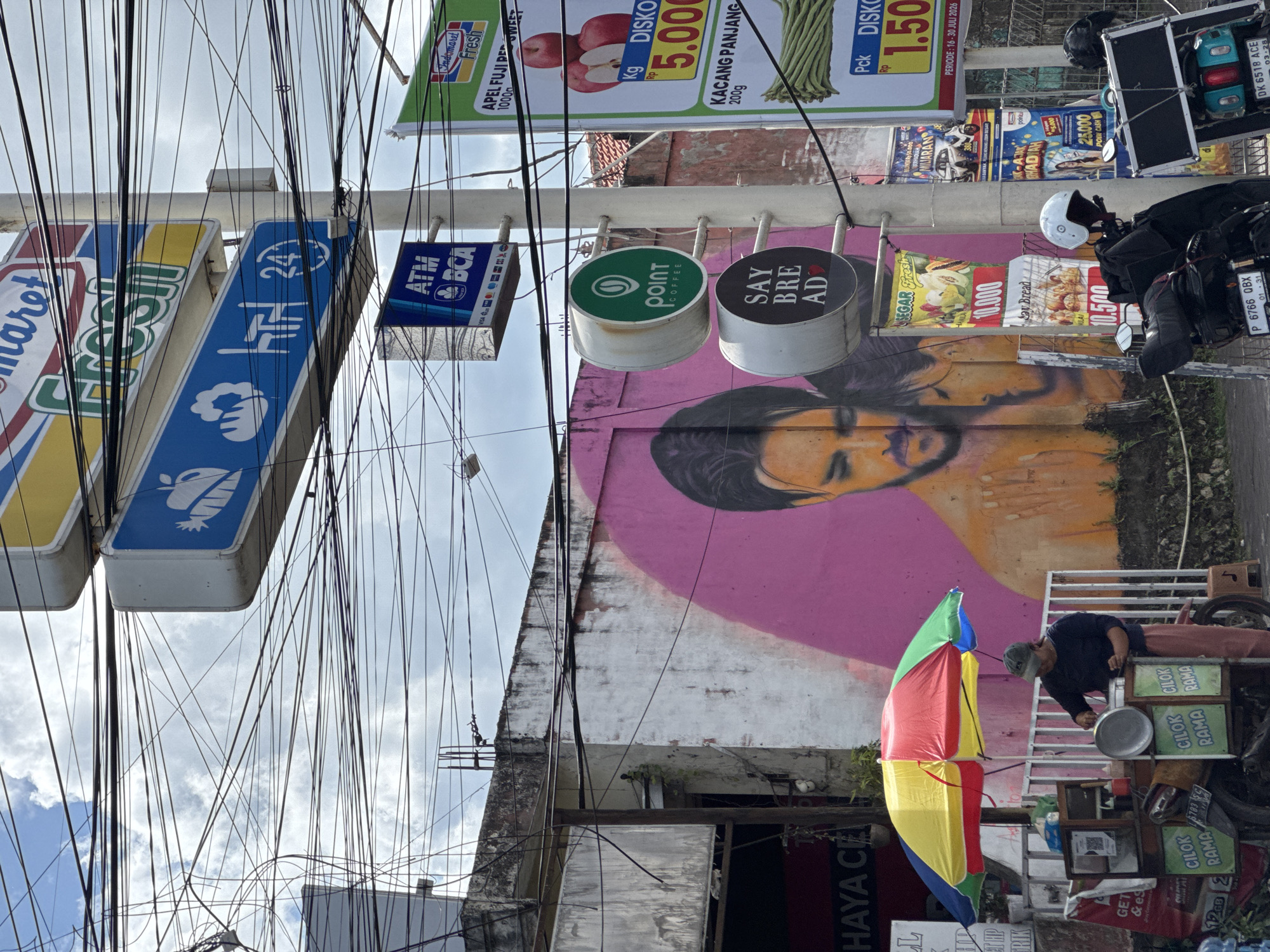 In Kuta Selatan, Indonesia, a street scene unfolds under a cloudy sky, dominated by a dense network of electrical wires overhead. A central utility pole displays a series of signs for "Indomaret," indicating a local convenience store chain. These signs advertise items like "Fresh," "ATM BCA," and promotions for "APEL FUJI RED SWEET" and "KACANG PANJANG." Below these, circular signs for "POINT COFFEE" and "SAY BREAD" are attached to the pole. To the left, a vibrant mural of a couple's faces adorns a pink wall. In the foreground, a person in a cap and dark shirt is tending to a street food cart beneath a colorful umbrella. The cart, decorated with signs for "CILOK RAMA," is laden with food items and a motorcycle. Another motorcycle, equipped with a black storage box, is parked nearby, with a white helmet resting on it. Signs and posters, including one for "Segar Fresh" and another with a car advertisement, are visible on the wall and utility pole.