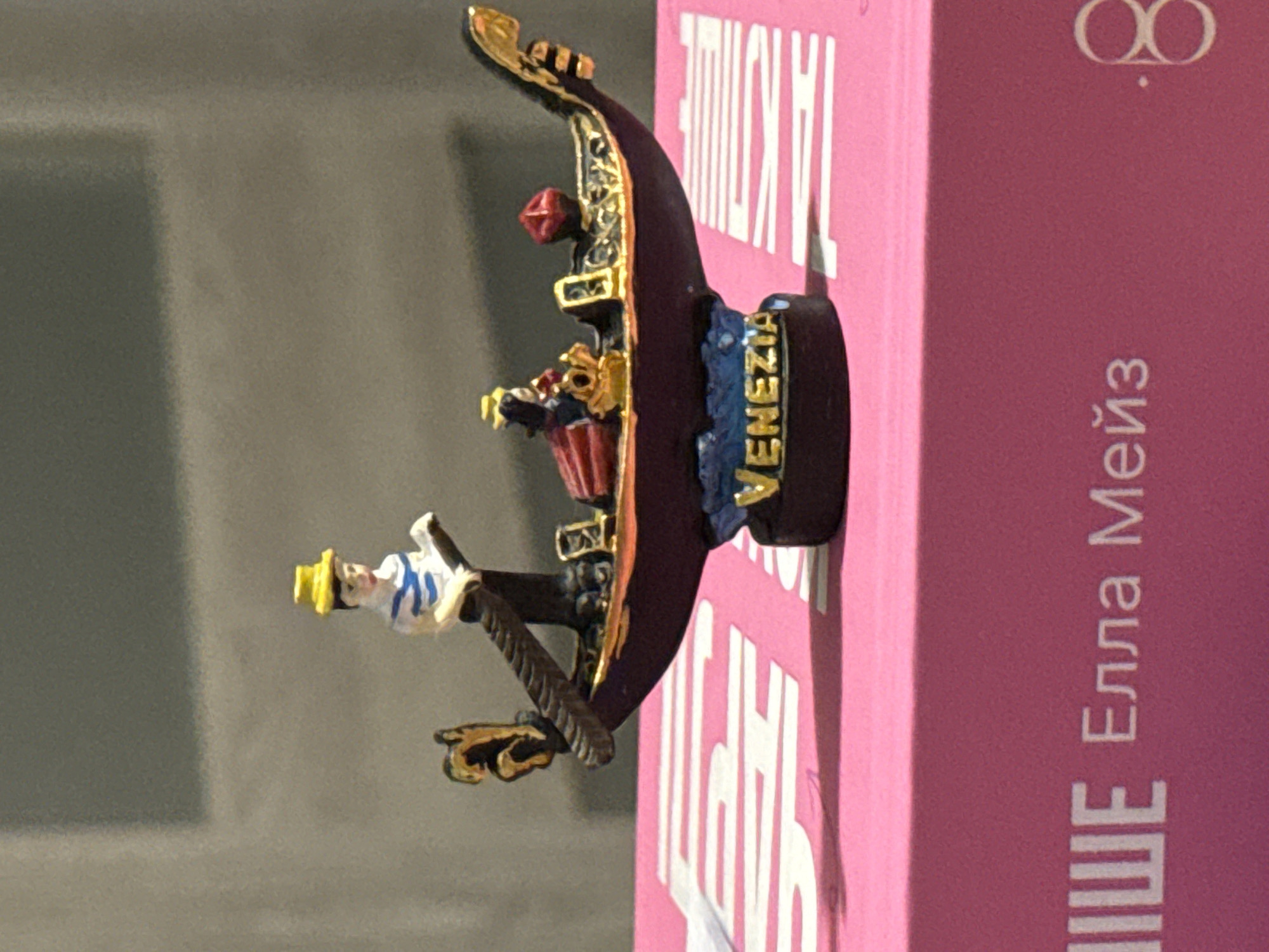 The image shows a miniature Venetian gondola souvenir with two figures, a gondolier and a passenger, positioned on a pink surface that appears to be the lid of a box. The gondolier wears a yellow hat and a blue and white striped shirt. The passenger is seated and wears a red garment. The gondola is dark red with gold trim. The word "VENEZIA" is written on a blue base below the gondola, which rests on the pink surface. The background is a blurred grey wall. The pink surface has white lettering on it, partially visible, which appears to be in Russian or a Cyrillic script. There is no indication of the city of Łódź, Poland, within the image itself.