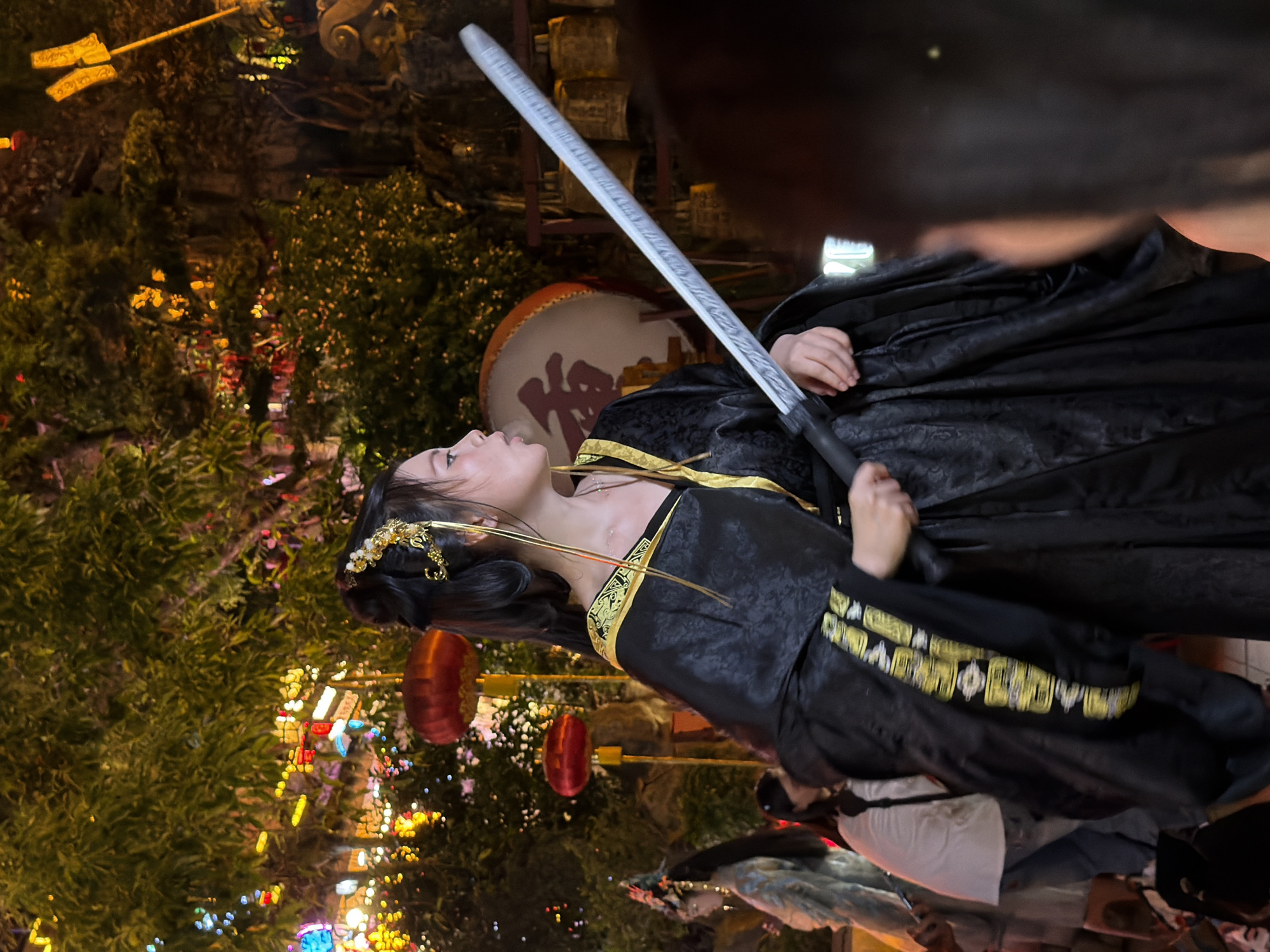A young East Asian woman, dressed in a black and gold traditional Chinese robe, stands holding a silver sword in her right hand. Her hair is styled up with gold ornaments. She is facing to her left, looking in that direction. Behind her, a large drum with Chinese characters is visible, along with a rack of bells. The background is filled with greenery, illuminated by colorful lights and red lanterns, suggesting an outdoor night setting. In the lower right of the image, the back of another person's head is visible, partially obscuring the woman. The scene appears to be in Yuzhong District, China, likely at a festival or cultural event.