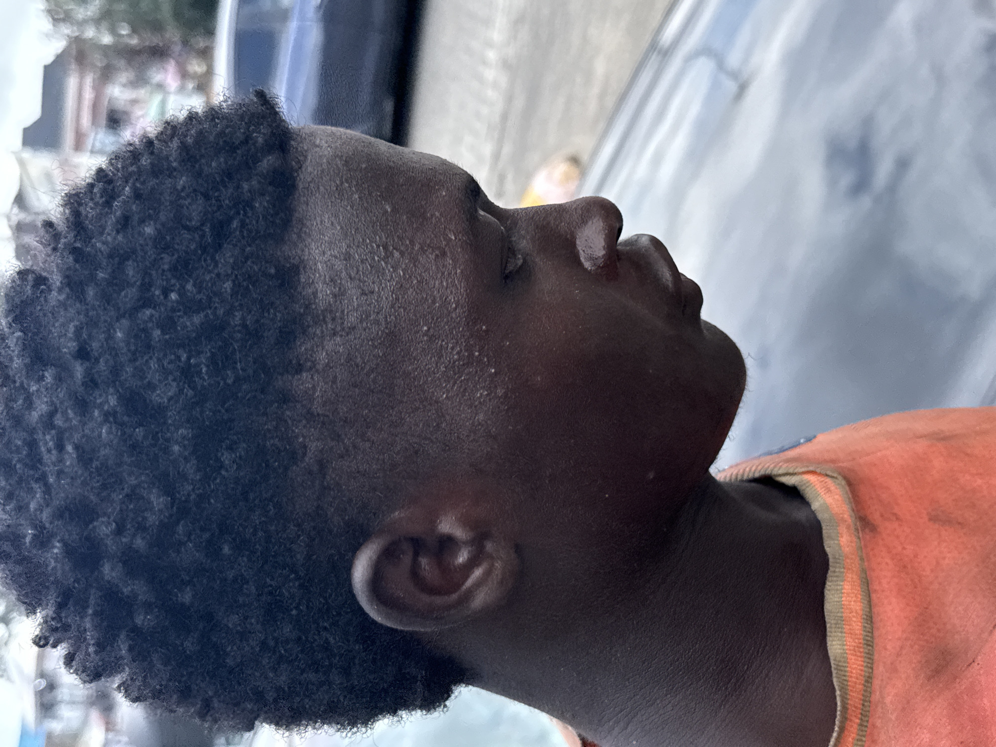 A medium close-up, tilted headshot of a Black man with a short Afro haircut and wearing a peach-colored t-shirt with two vertical green and tan stripes along the collar. He is gazing upward and slightly to the right. The background appears to be urban, with a car and indeterminate buildings. Based on the prompt specifying "Ado, Nigeria," the location is within that city.