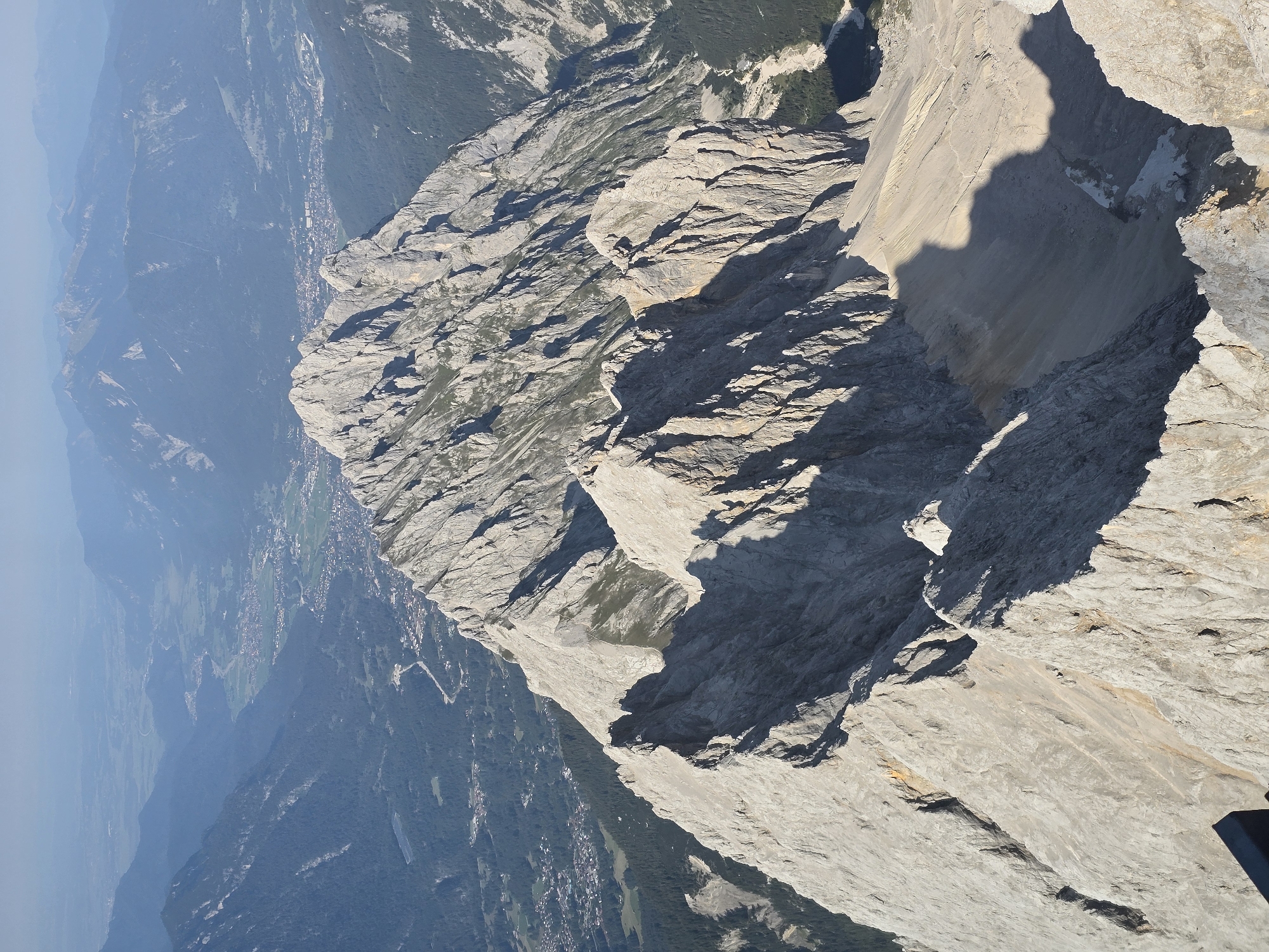 This is an aerial, eye-level, full shot of a rugged, mountainous landscape with a town nestled in the valley below. The foreground is dominated by sharp, gray, rocky peaks and ridges, indicative of a high alpine environment. Sunlight casts dramatic shadows across the scree slopes and rock formations. In the mid-ground, a prominent mountain range with jagged peaks stretches across the frame. Further in the distance, a hazy valley unfolds, revealing a town with scattered buildings, roads, and a river, suggesting the presence of human settlement in Grainau, Germany. Beyond the valley, more mountain ranges recede into the blue haze of the horizon under a clear, bright sky. There are no people or discernible actions taking place in the image; it is a static, natural landscape with a town visible in the distance.